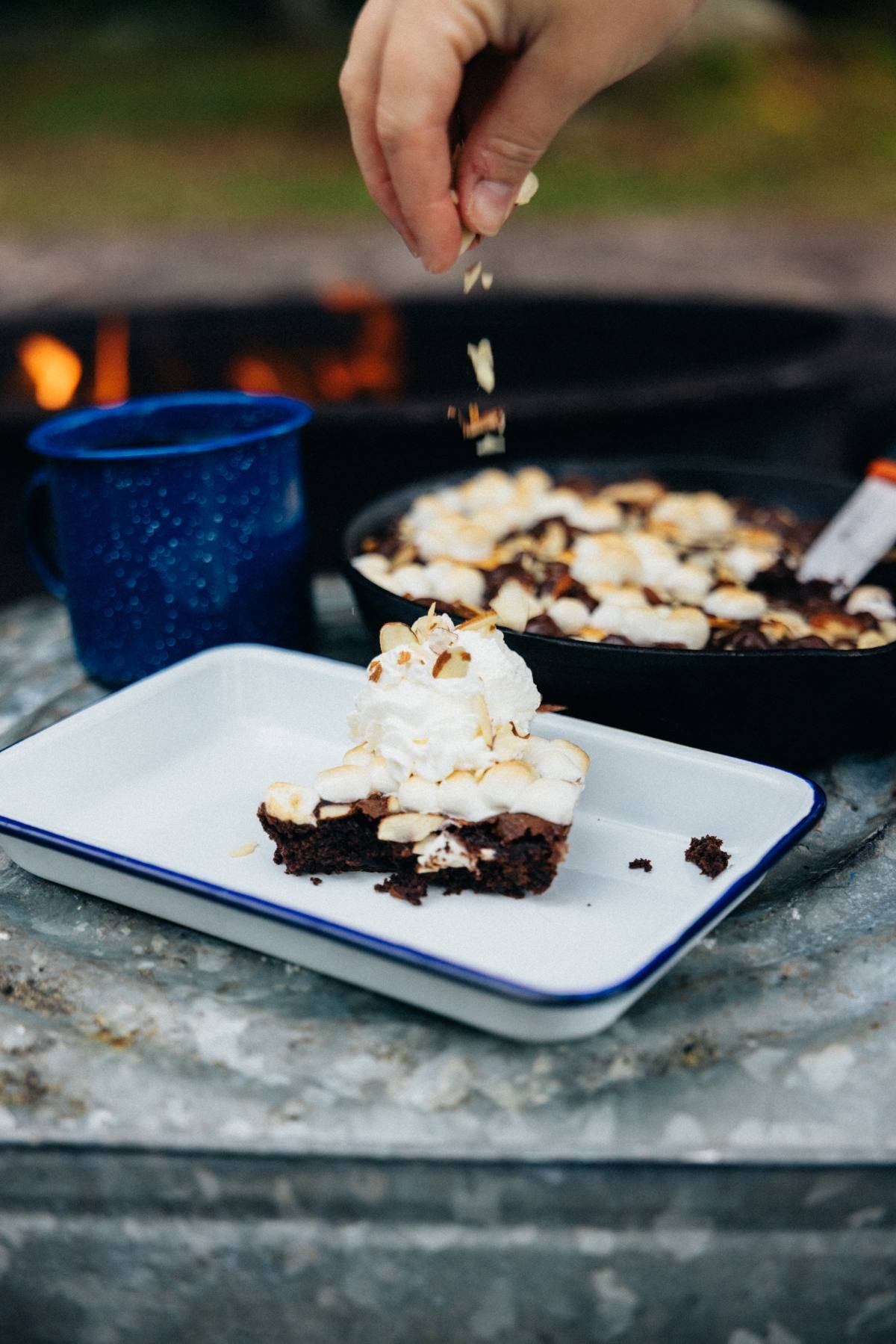 A hand sprinkles nuts onto a brownie with ice cream; coffee and dessert pan are in the background.