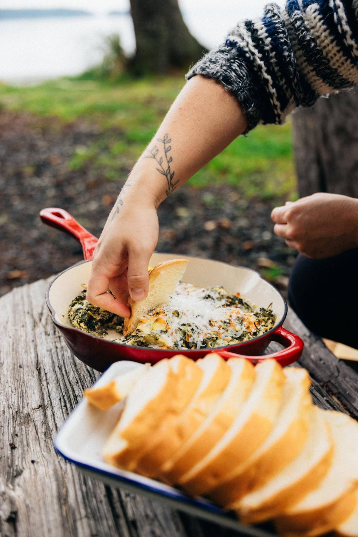 A hand dips bread into a skillet of cheesy spinach dip outdoors, with sliced bread in the foreground.