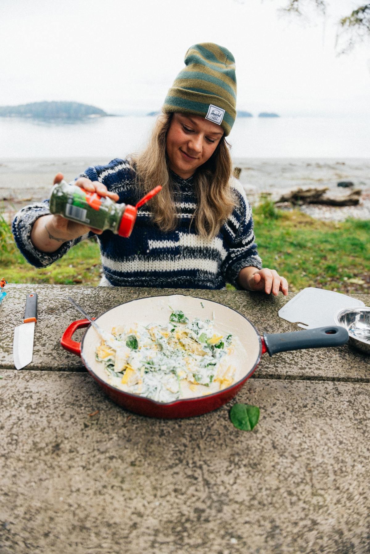 A woman in a striped sweater seasons food in a pan outdoors near a beach.