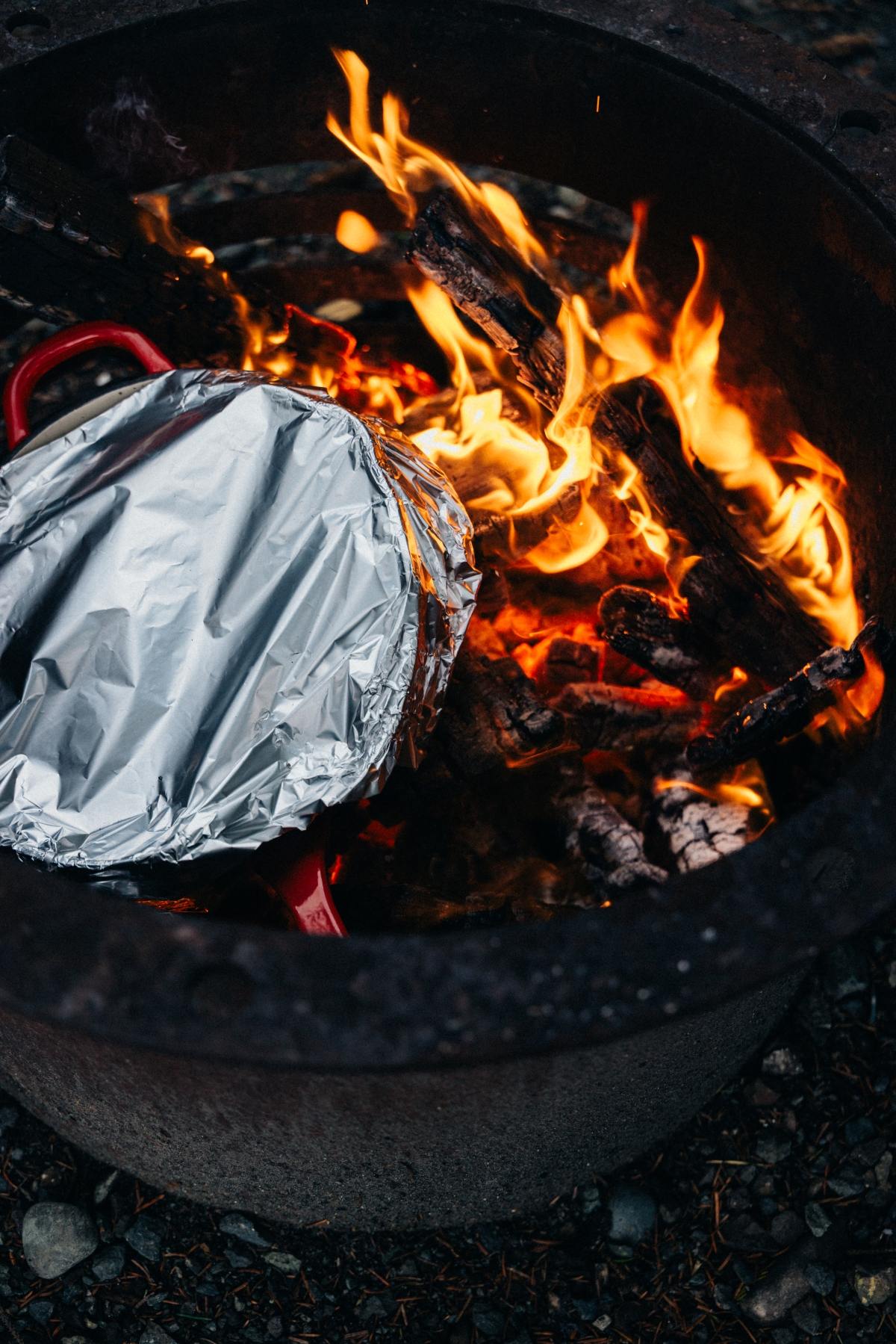 A foil-covered pot sits over burning wood in a fire pit, surrounded by bright orange flames.
