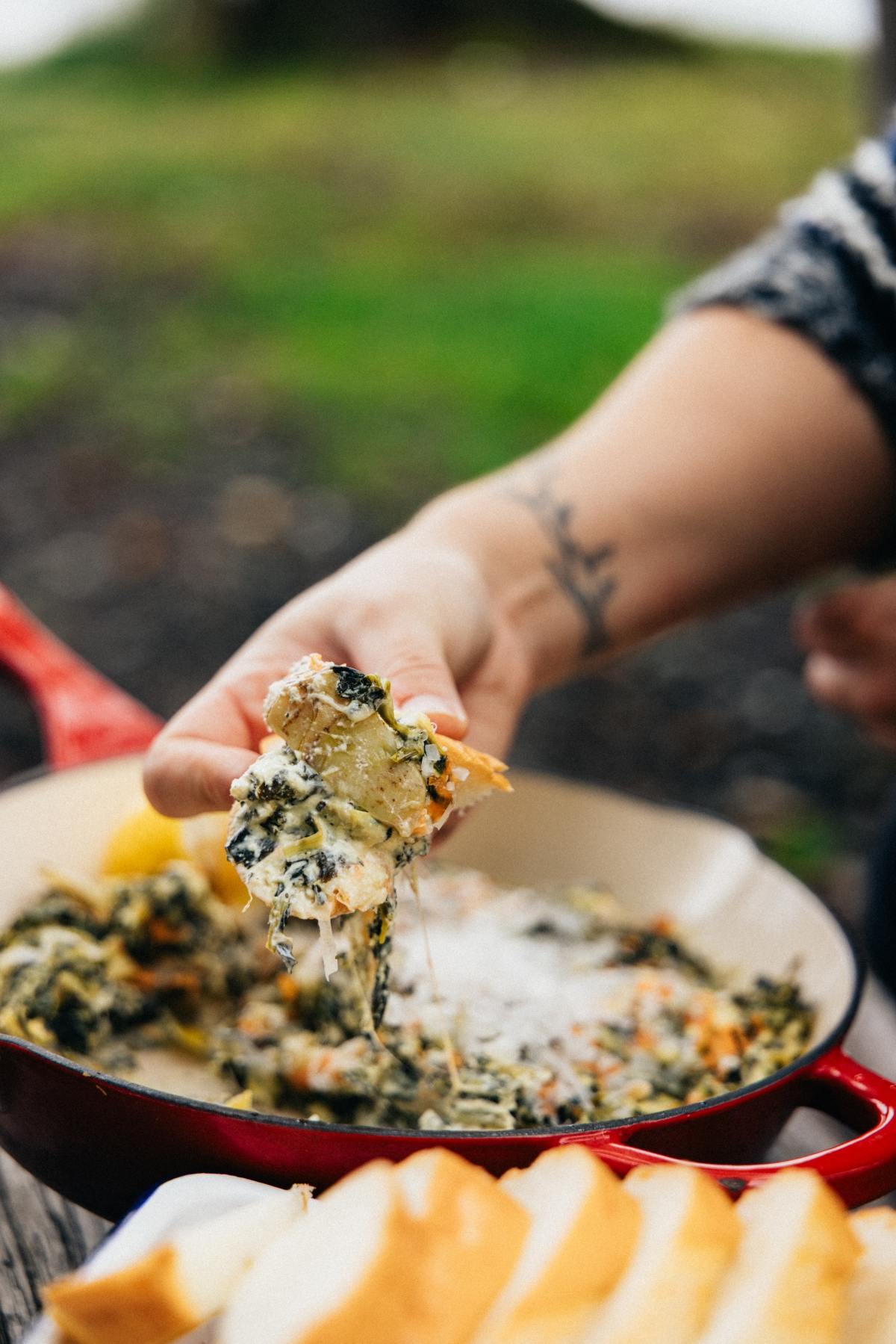 A hand scoops cheesy spinach dip from a skillet, with slices of bread in the foreground.