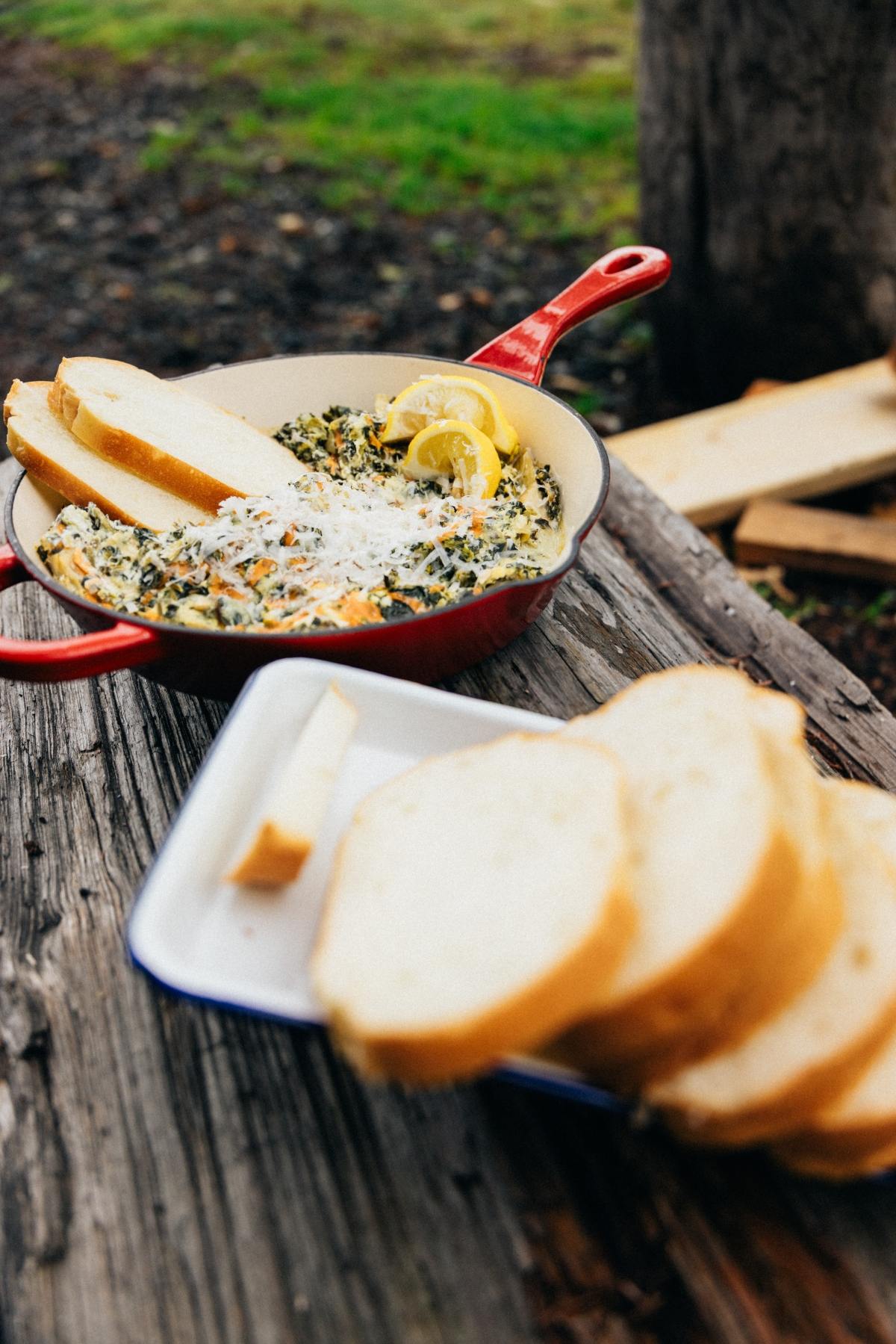 A skillet of creamy spinach dip with lemon and sliced bread, set on a rustic wooden table outdoors.