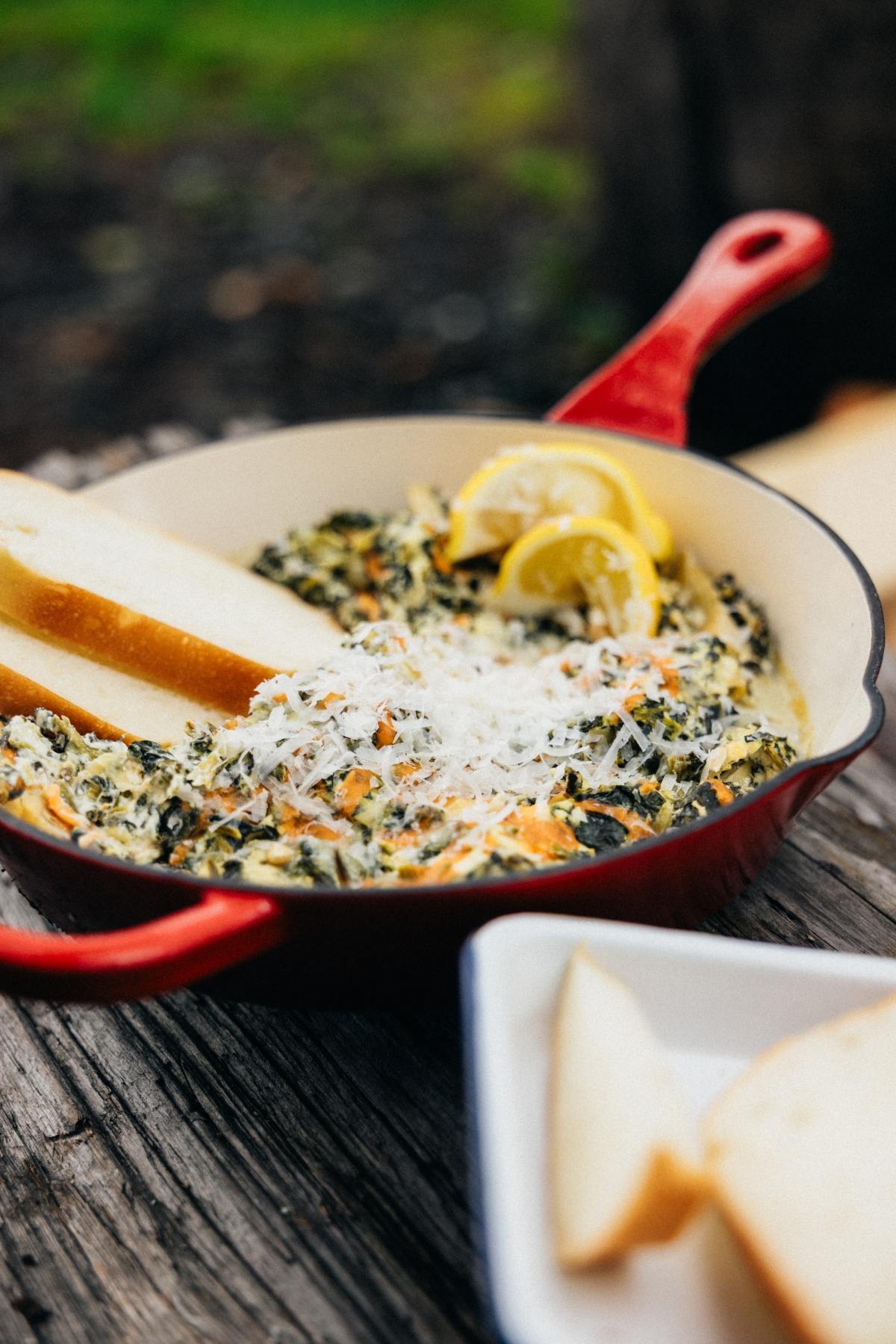 Red skillet with spinach dip, shredded cheese, lemon slices, and bread slices on a rustic wooden surface.