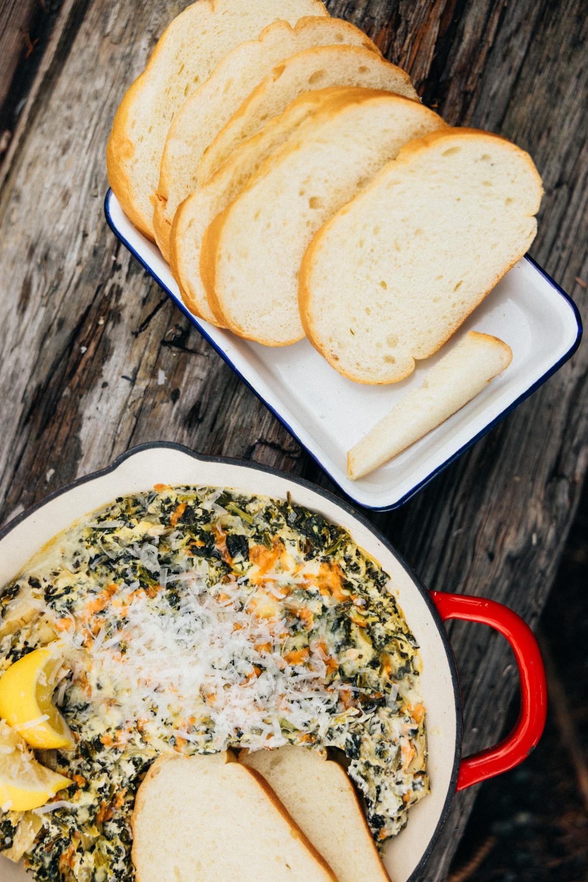 A dish of creamy spinach dip with lemon slices, served with sliced white bread on a tray.