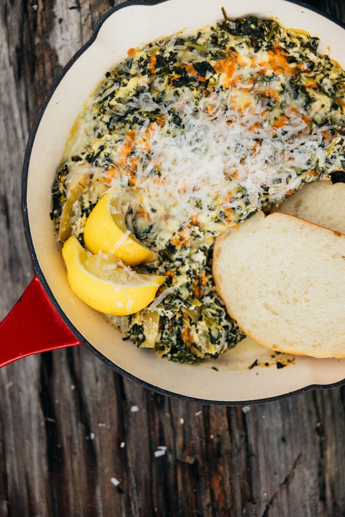 Creamy spinach dip in a skillet, topped with cheese, lemon wedges, and served with slices of bread.