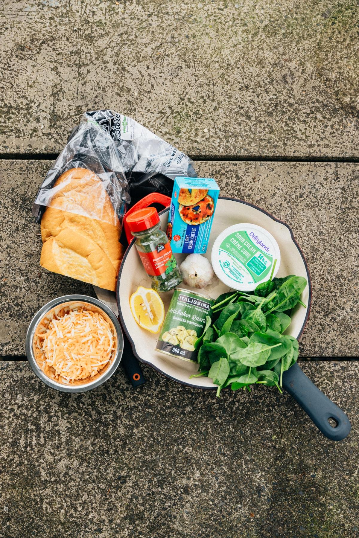 A skillet with bread, cheese, spinach, lemon, cream cheese, garlic, and seasonings on a concrete surface.