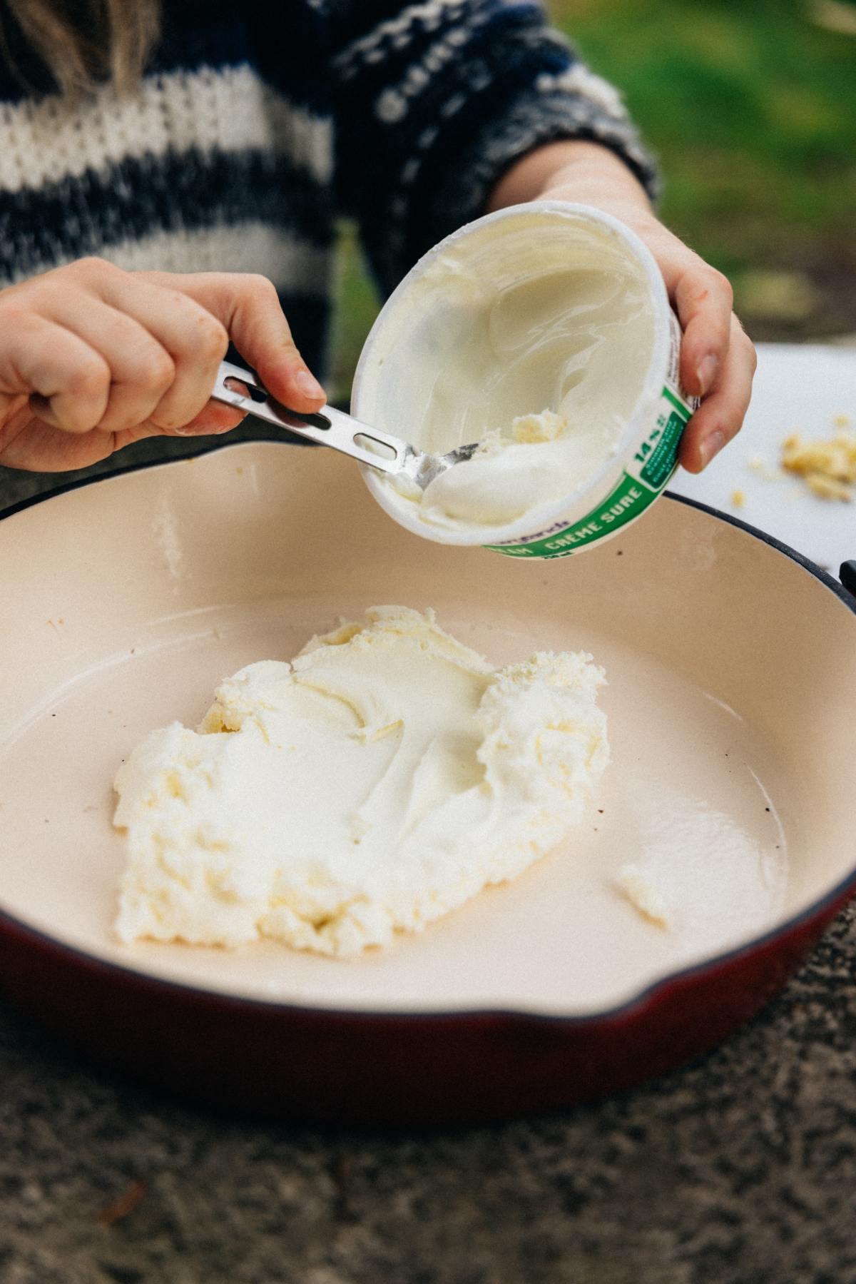 Person scooping white spread from container into a large cream-colored pan outdoors.