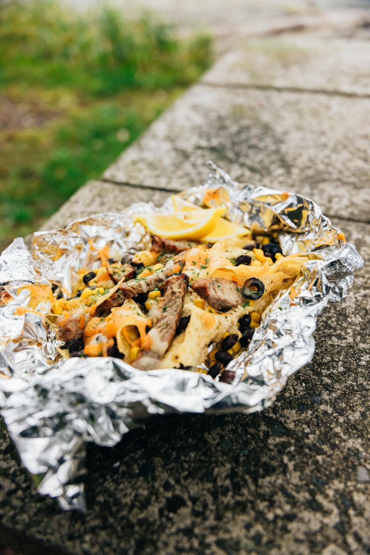 Nachos with grilled meat, cheese, olives, and beans in foil on a stone surface outdoors.