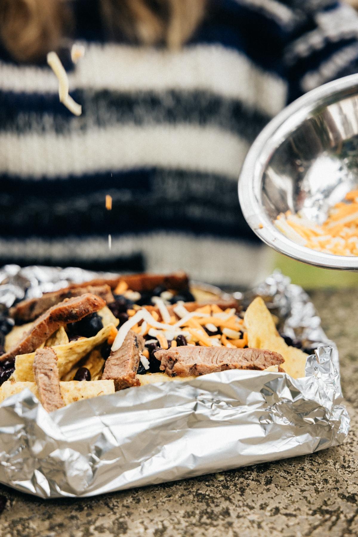 A person sprinkles shredded cheese onto nachos with steak on foil outdoors.