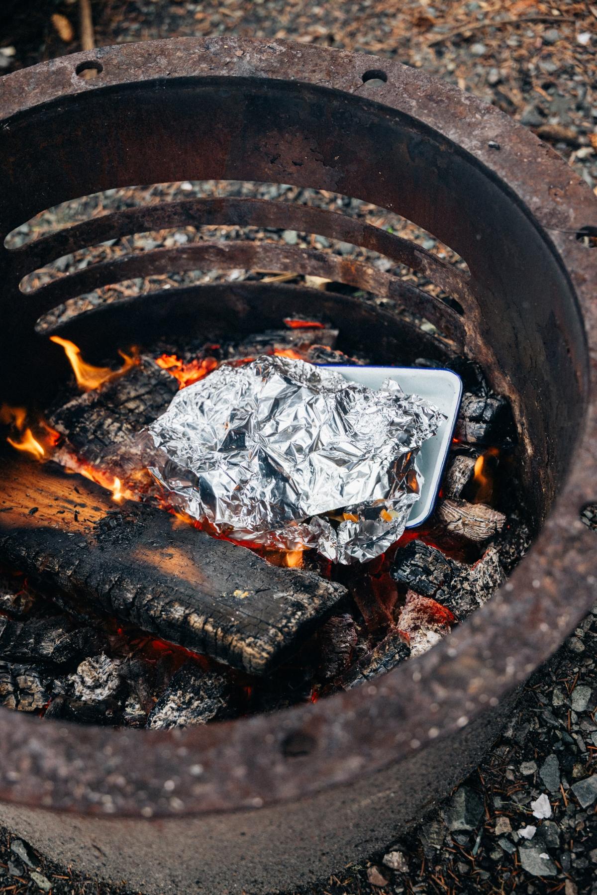 Aluminum foil-wrapped food cooking on a tray over a campfire in a metal fire ring.