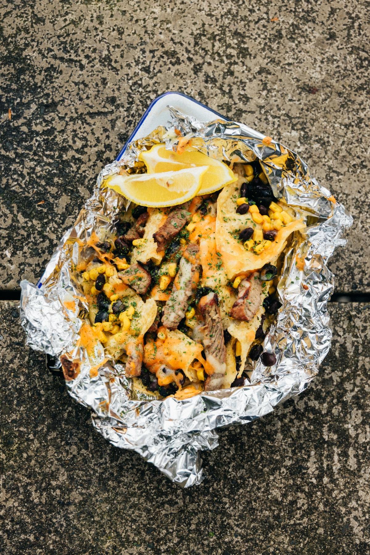 A foil tray with loaded nachos, beef, cheese, black beans, and lemon wedges on a textured surface.