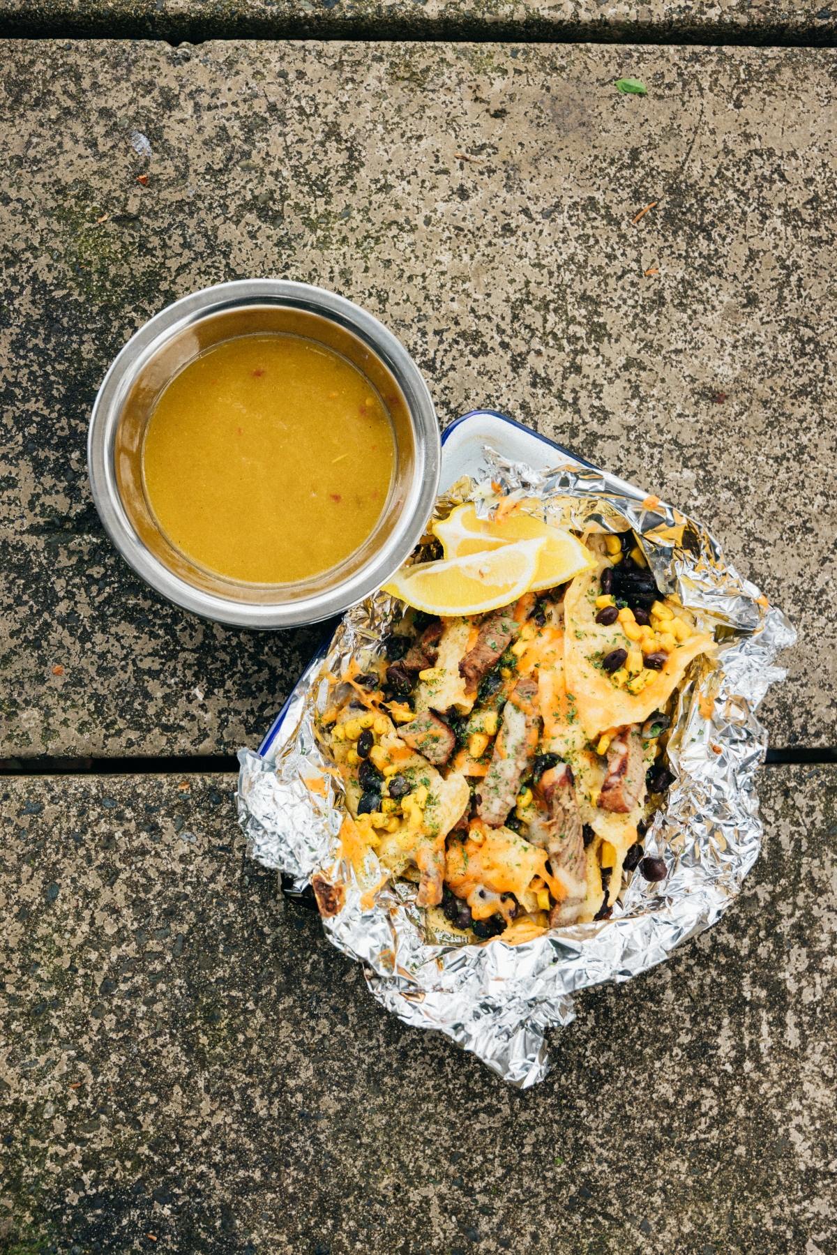 Bowl of soup and foil tray of nachos with cheese, beans, and lemon slices on a concrete surface.