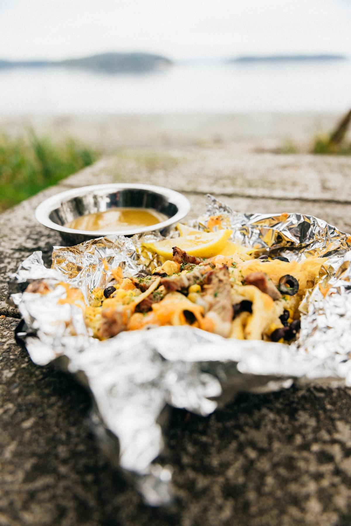 Foil-wrapped nachos with cheese and olives on a stone table, sauce in a metal dish, outdoors by the water.