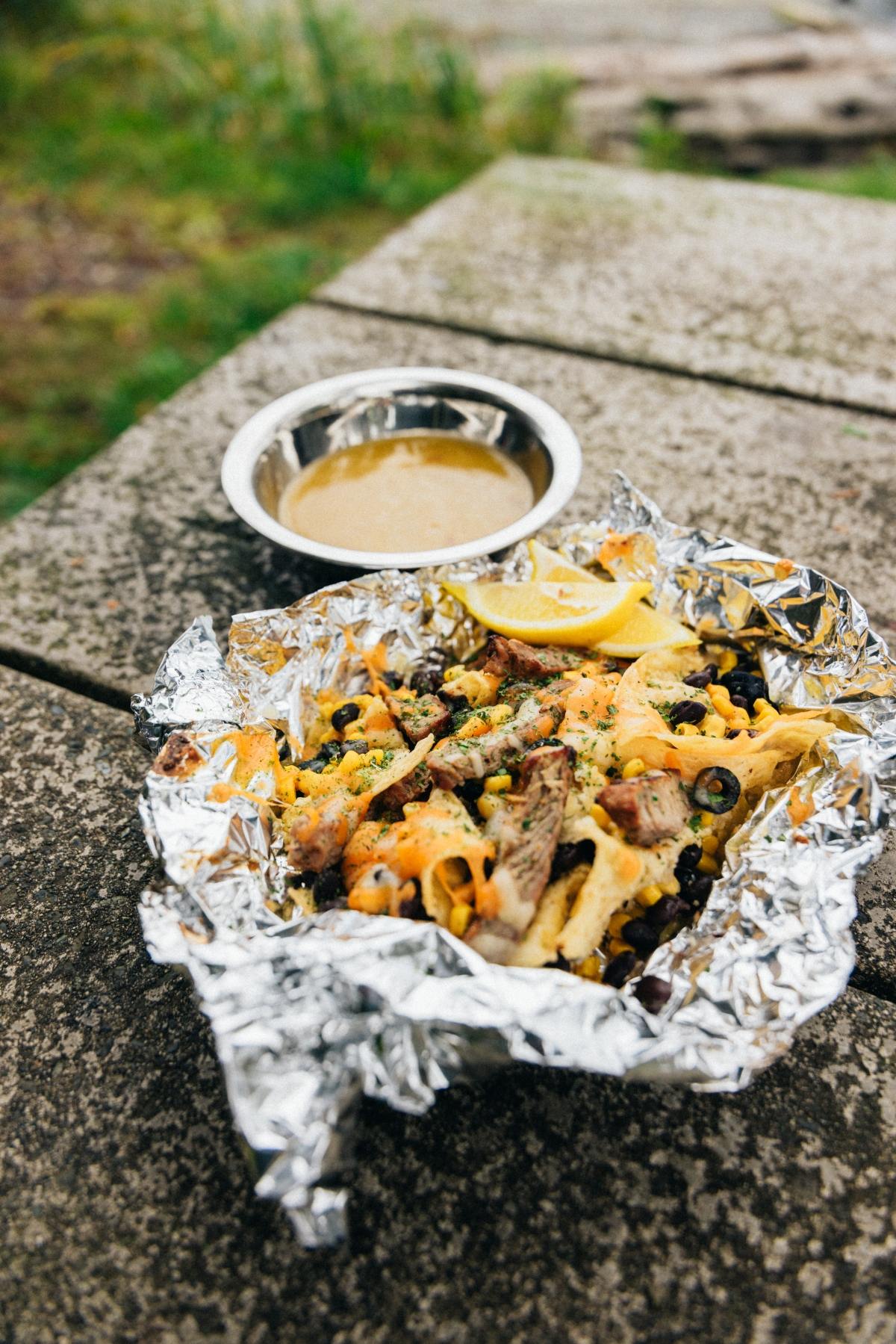 Waffle fries with toppings in foil, lemon wedge, and sauce in a metal dish on a picnic table outdoors.
