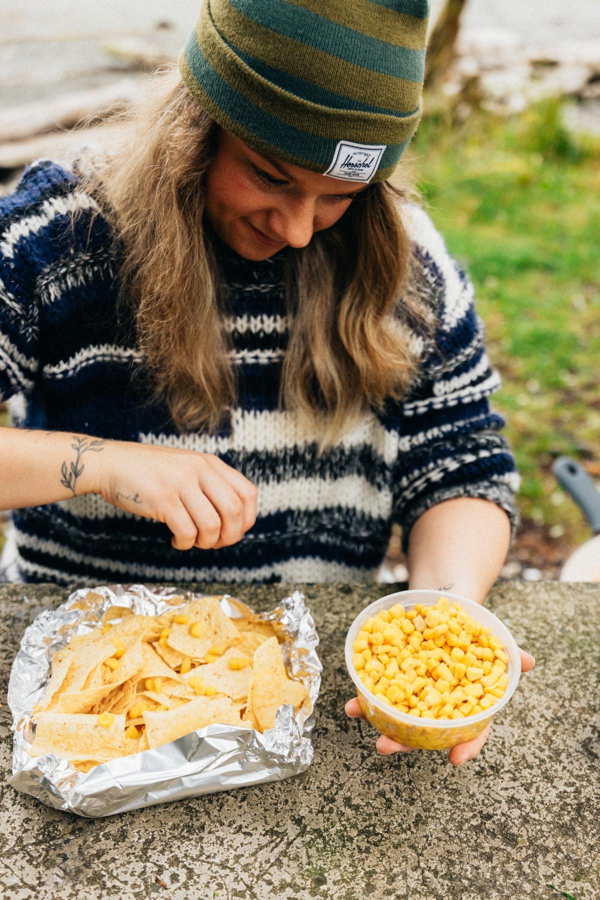 Person in a striped sweater and beanie adds corn to nachos outdoors on a stone surface.