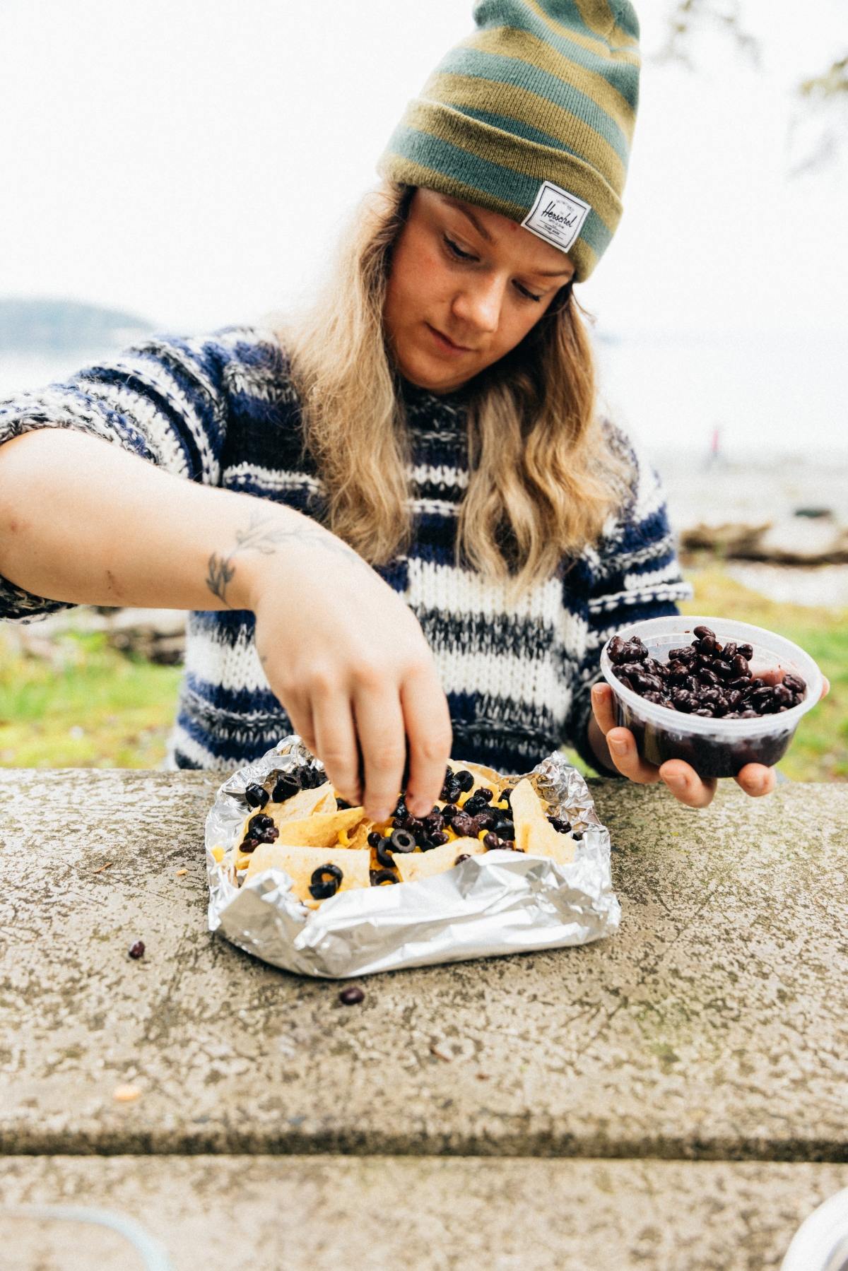 Woman in a beanie adds black beans to nachos on a picnic table outdoors.