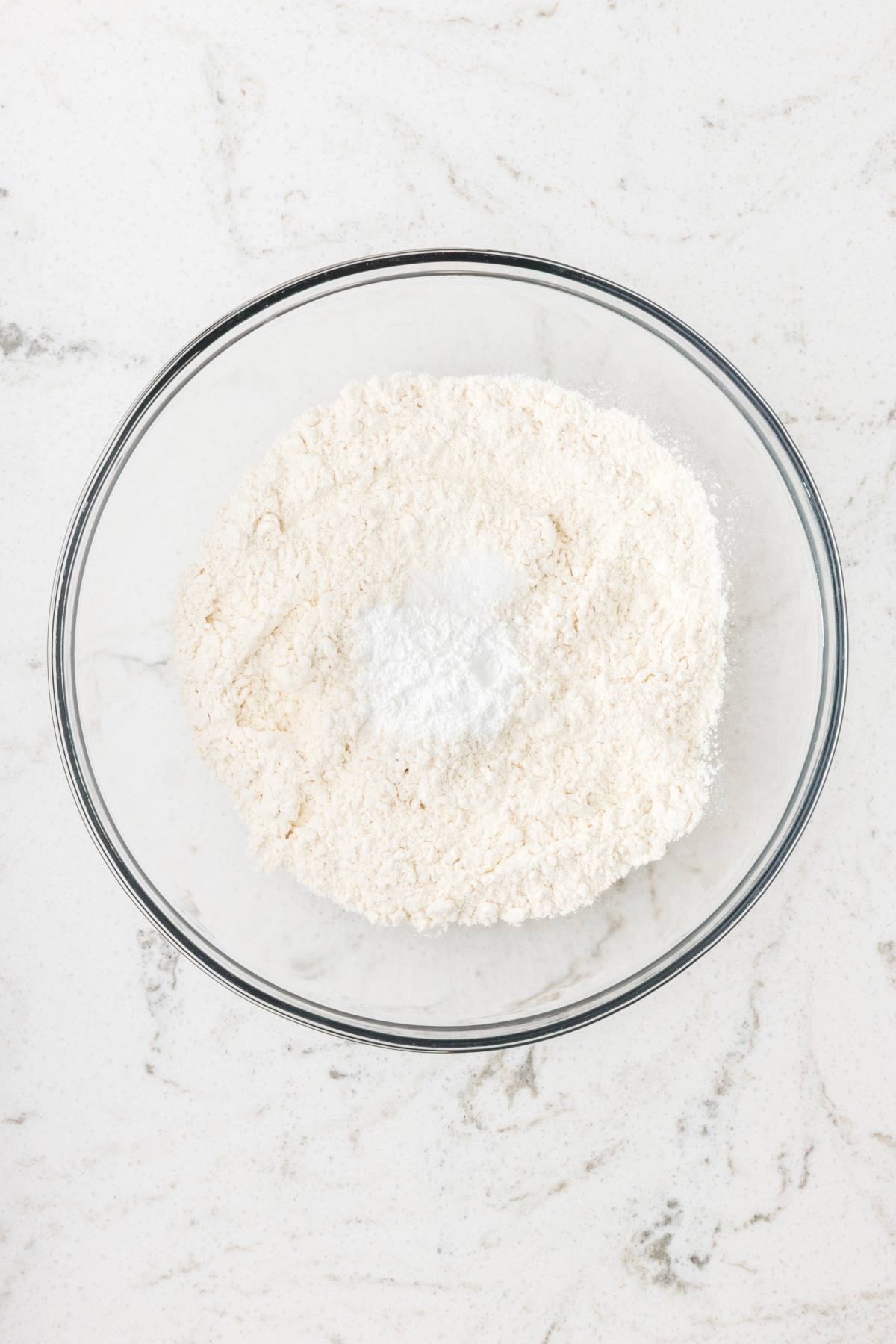 A glass bowl with flour and baking powder on a white marble countertop.