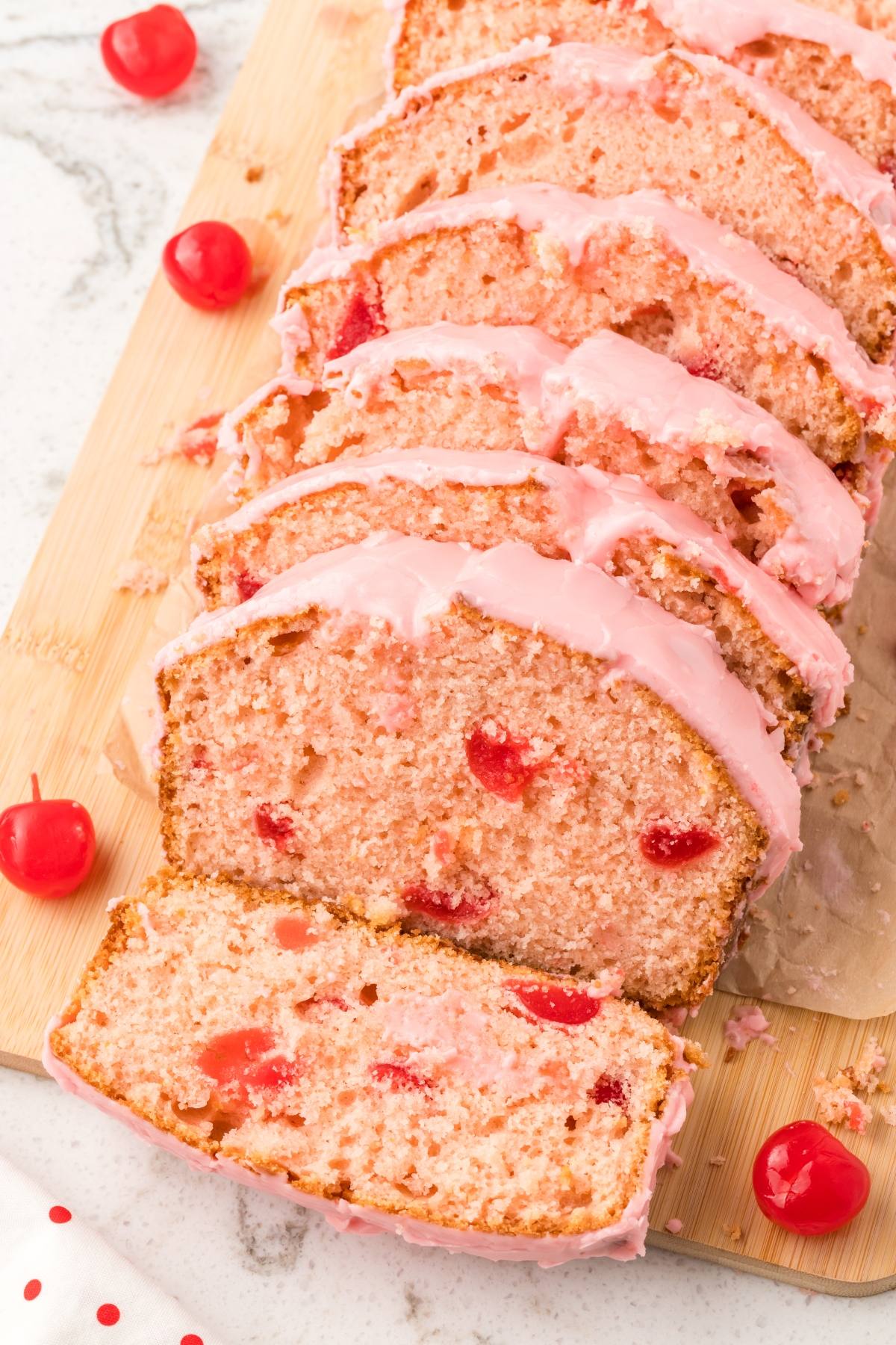 Sliced cherry bread loaf with pink icing and whole cherries on a wooden board.