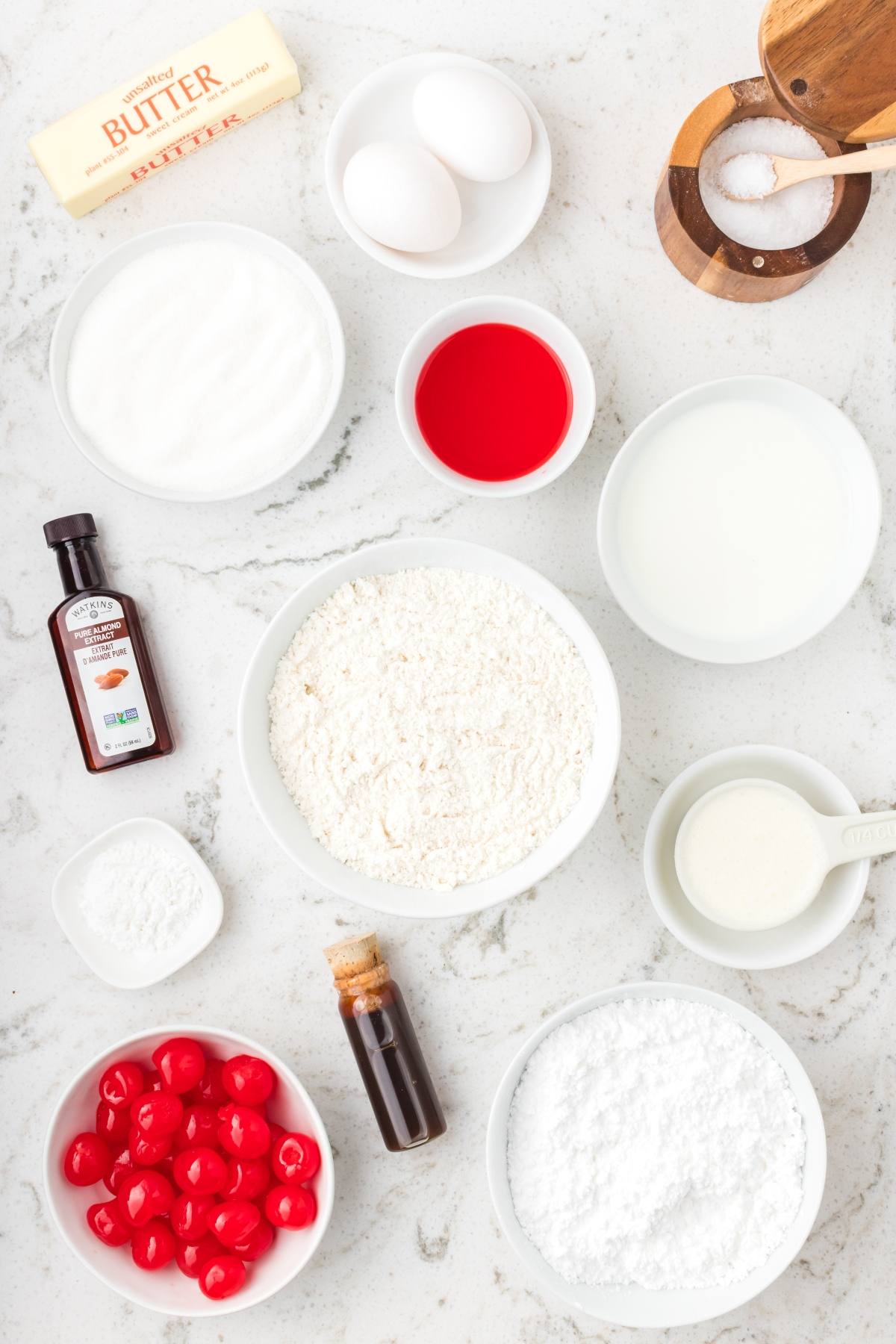 Baking ingredients on a counter: butter, eggs, sugar, flour, milk, cherries, vanilla, salt, and bowls of liquids.