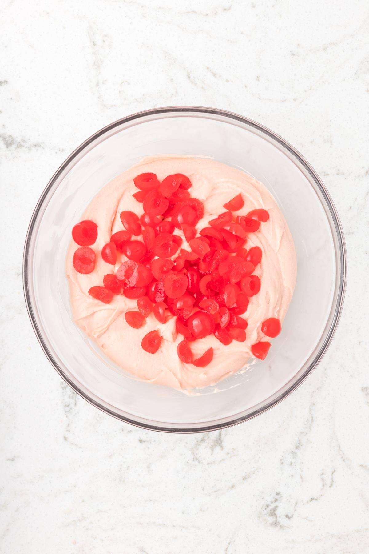 A glass bowl with pink whipped mixture topped with chopped red cherries on a white marble surface.