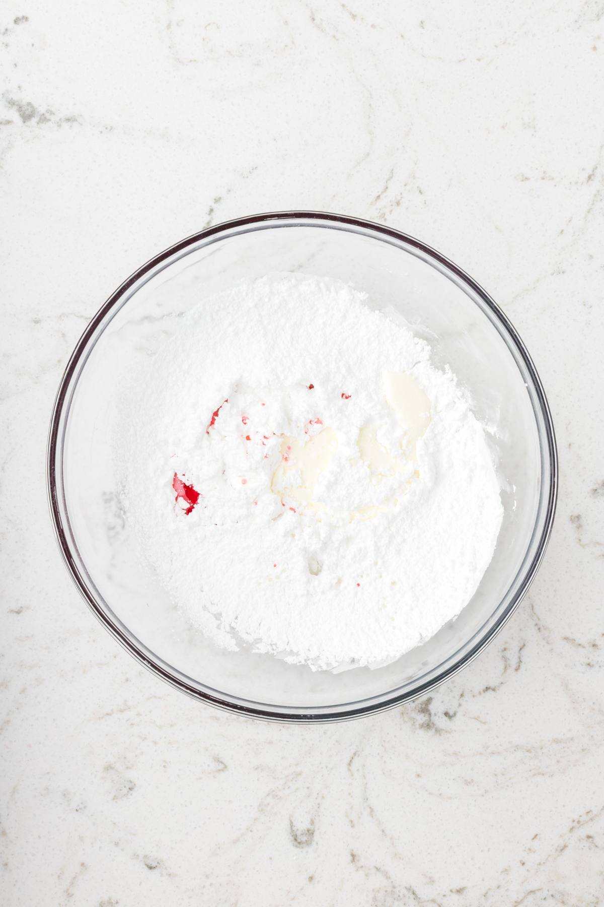 A glass bowl with powdered sugar, milk, and red food coloring on a white marble surface.