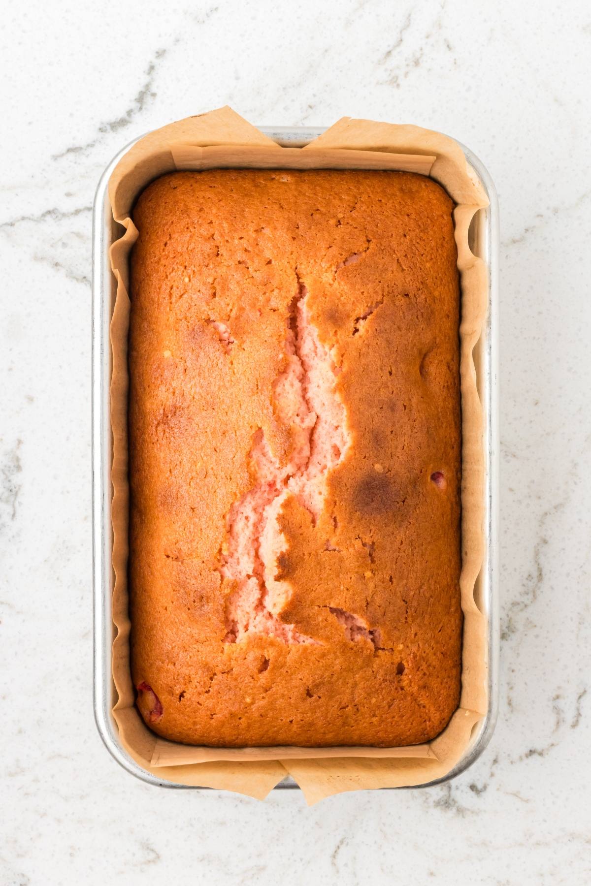 A loaf of golden-brown bread with a crack down the middle, in a parchment-lined baking pan on a marble surface.