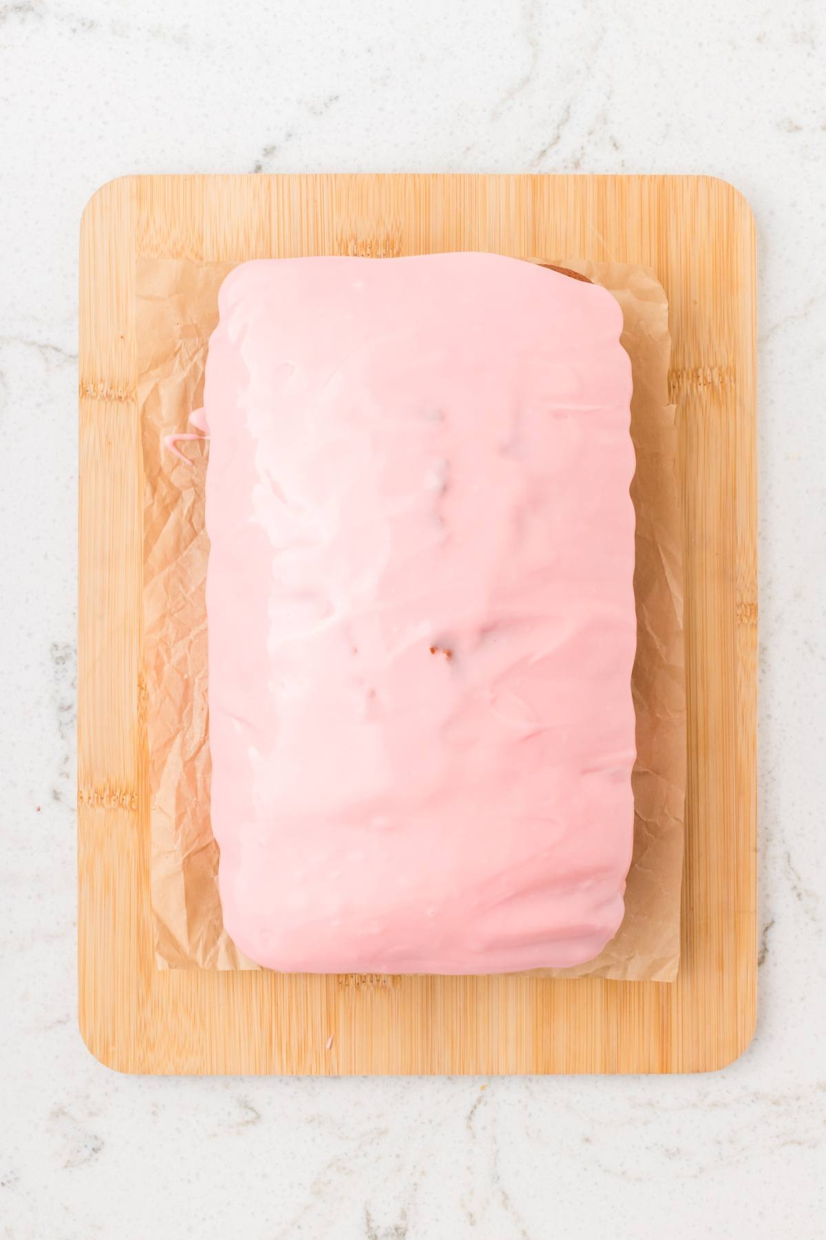 A loaf cake covered in smooth pink icing sits on parchment paper atop a wooden cutting board.