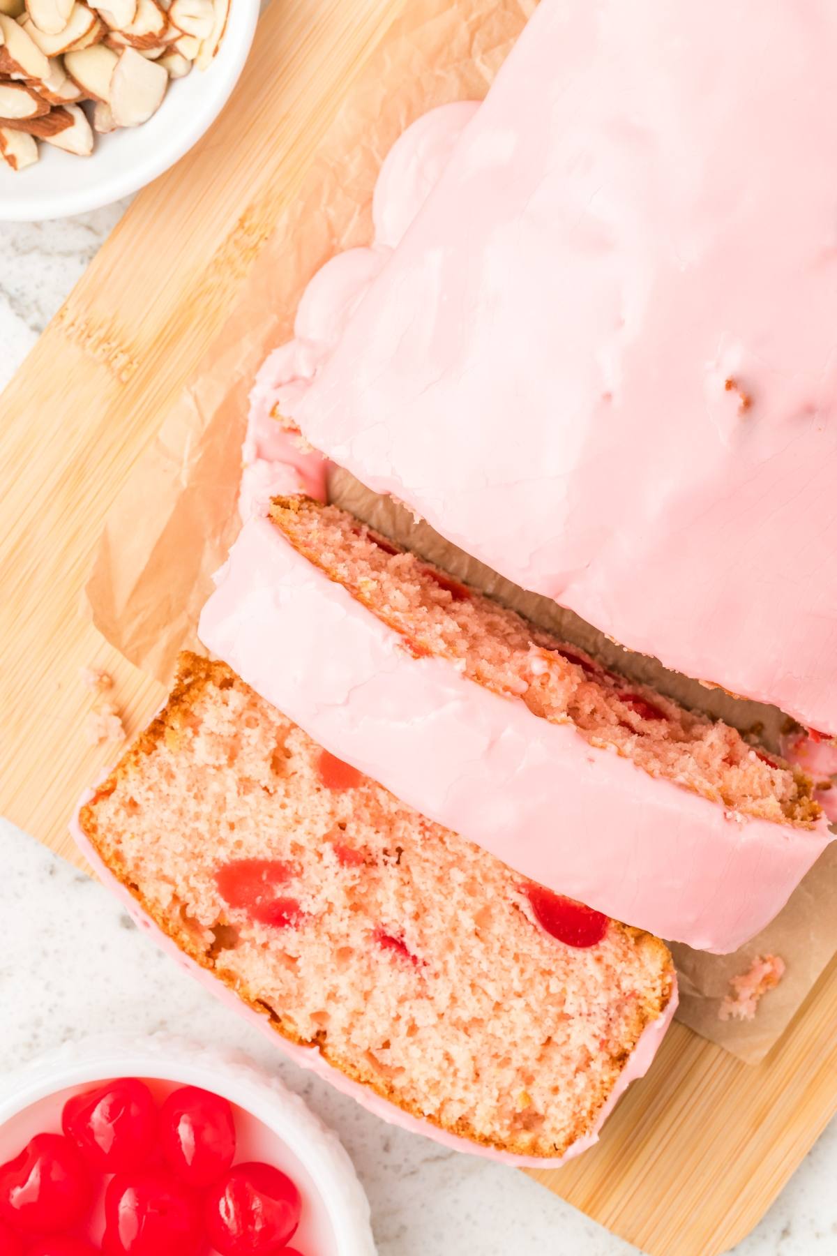 A sliced loaf of cherry bread with pink icing on a wooden board, next to a bowl of cherries.