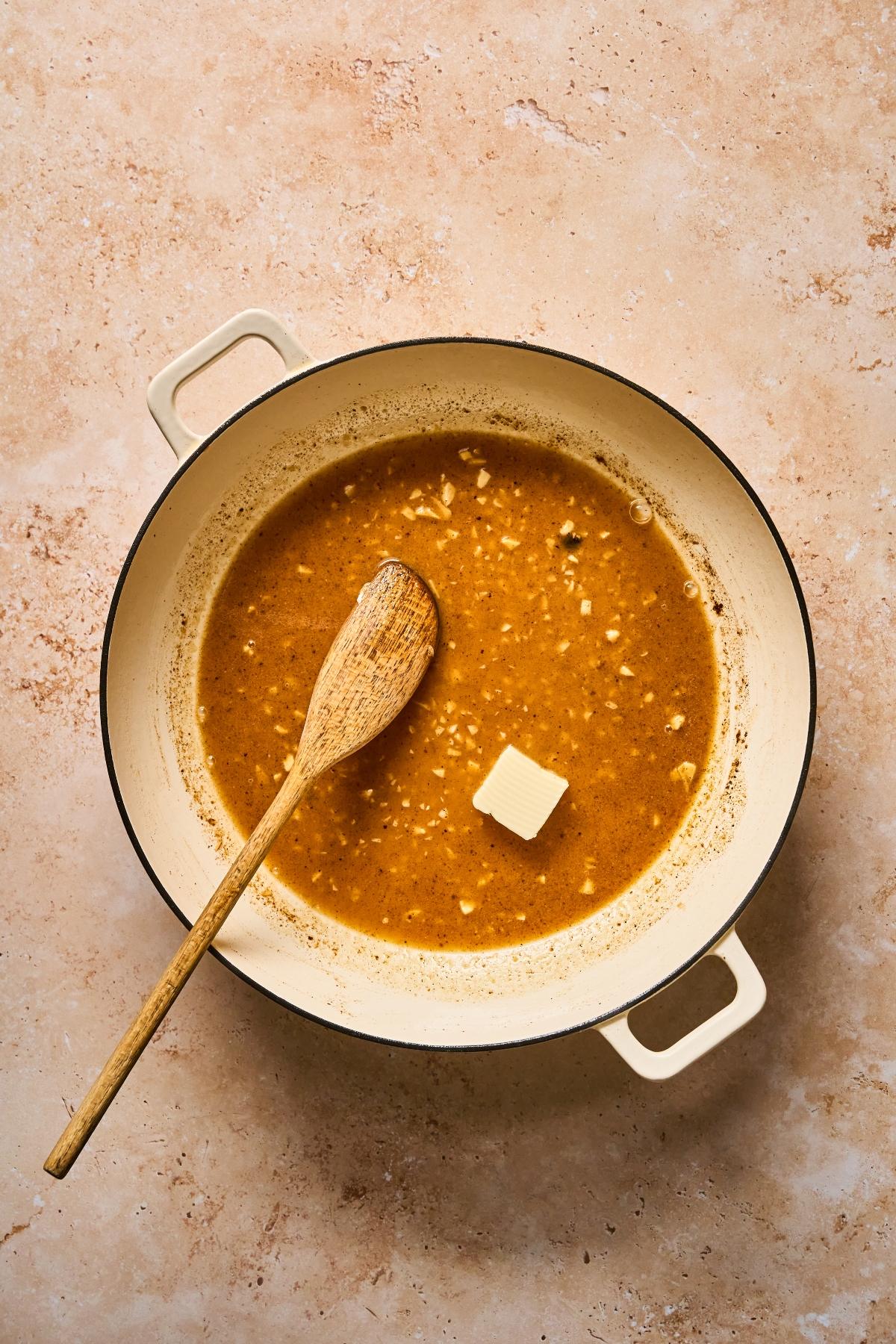 A white pot with brown sauce, a wooden spoon, and a pat of butter on a light countertop.