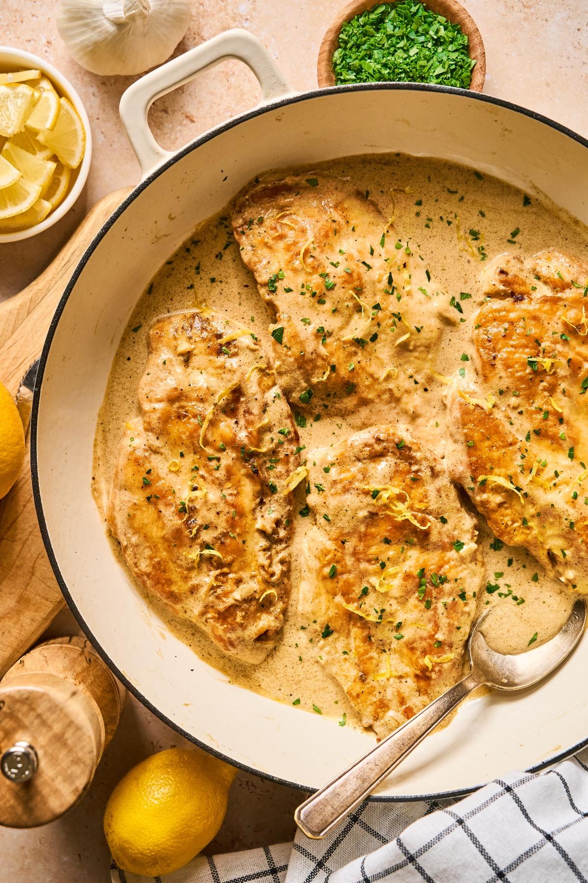 Chicken breasts in creamy sauce, garnished with herbs, in a pan with a spoon, lemons, and seasonings nearby.