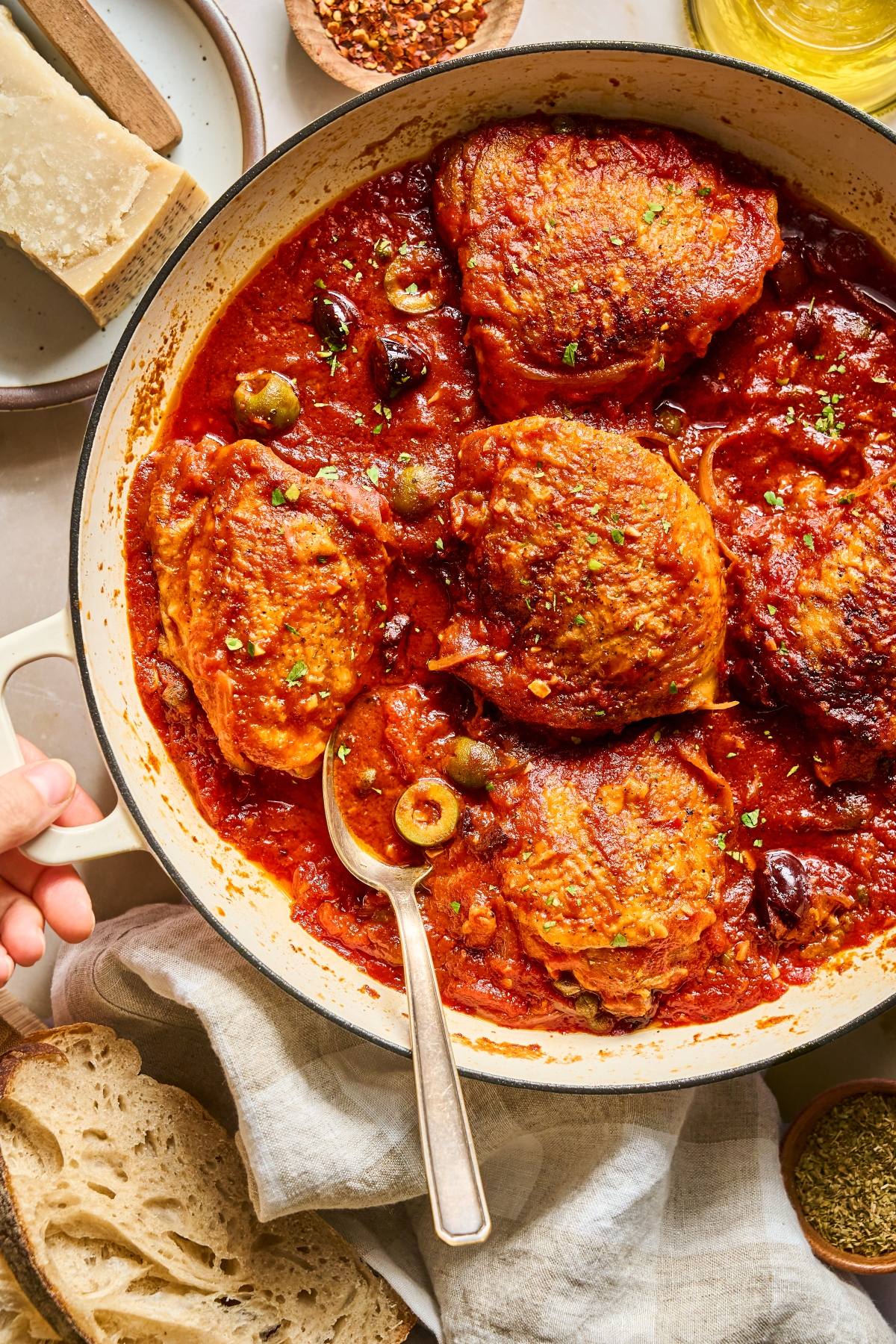 Chicken thighs in a red tomato sauce with olives in a pan, served with bread on the side.