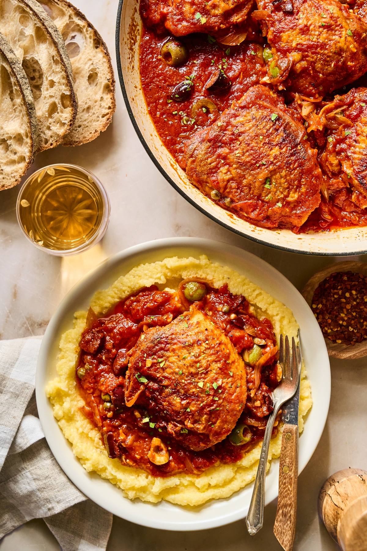 Plate of polenta topped with braised chicken in tomato sauce, with bread slices and a glass of white wine.
