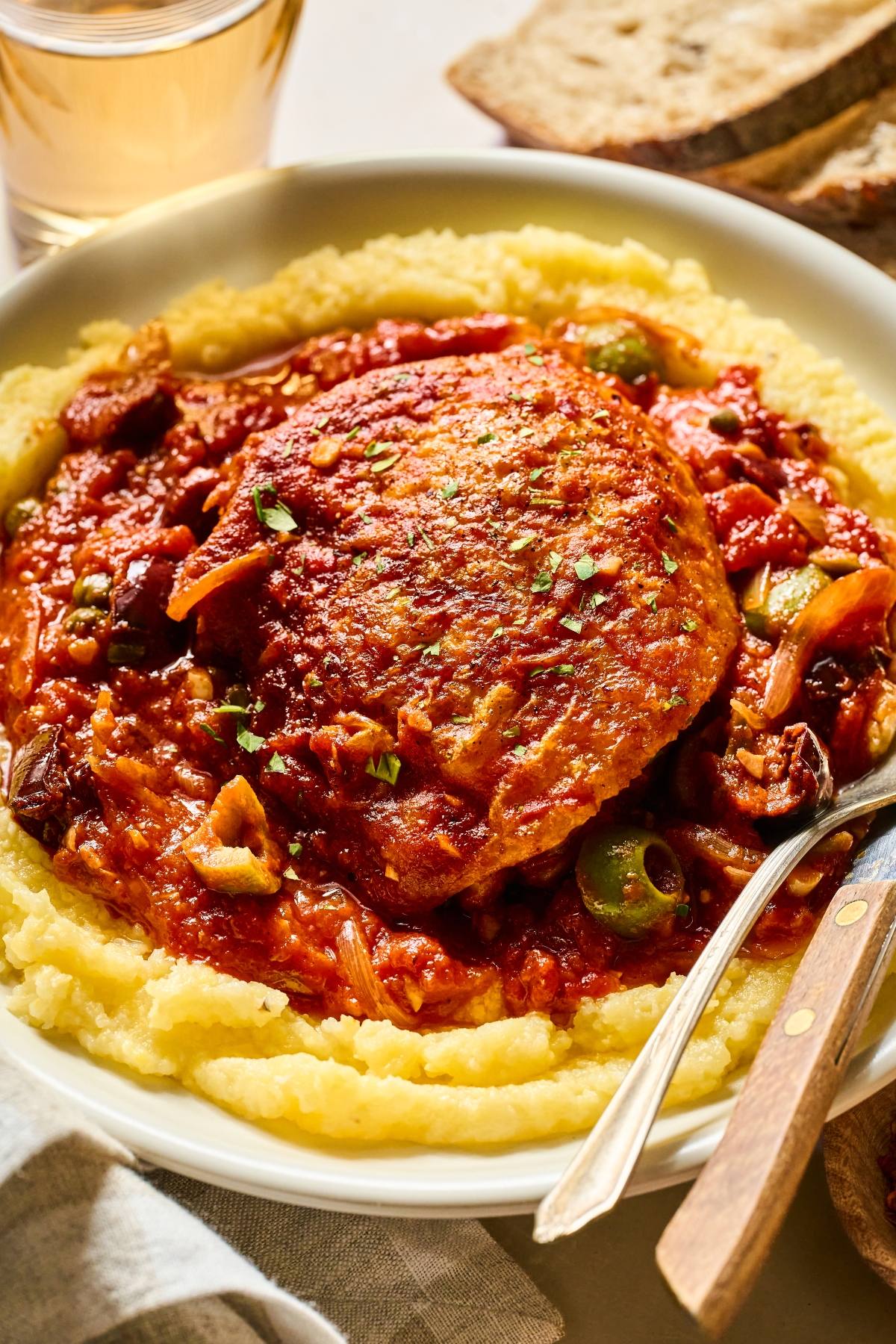 A plate of chicken puttanesca in tomato sauce over creamy polenta, served with bread and a glass of drink.
