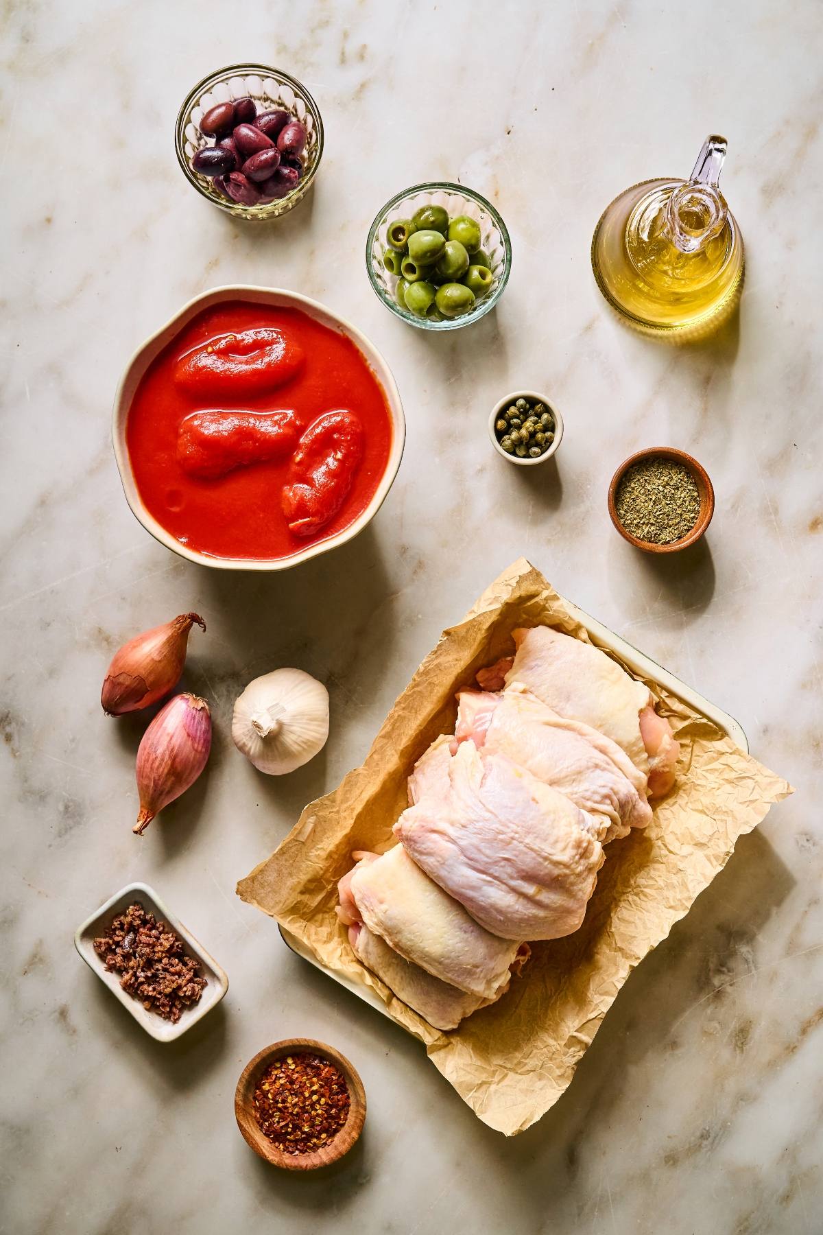 Raw chicken, tomatoes, olives, capers, olive oil, garlic, shallots, spices, and seasonings on a marble surface.