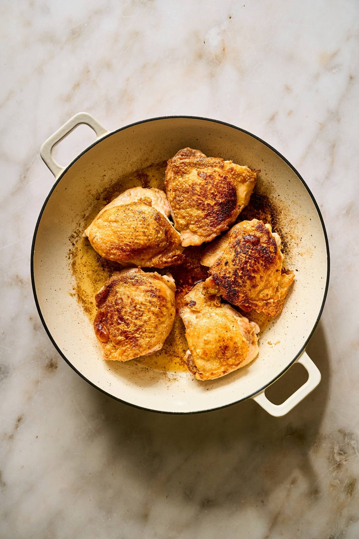 Four browned chicken thighs cooking in a round, cream-colored pot on a marble surface.