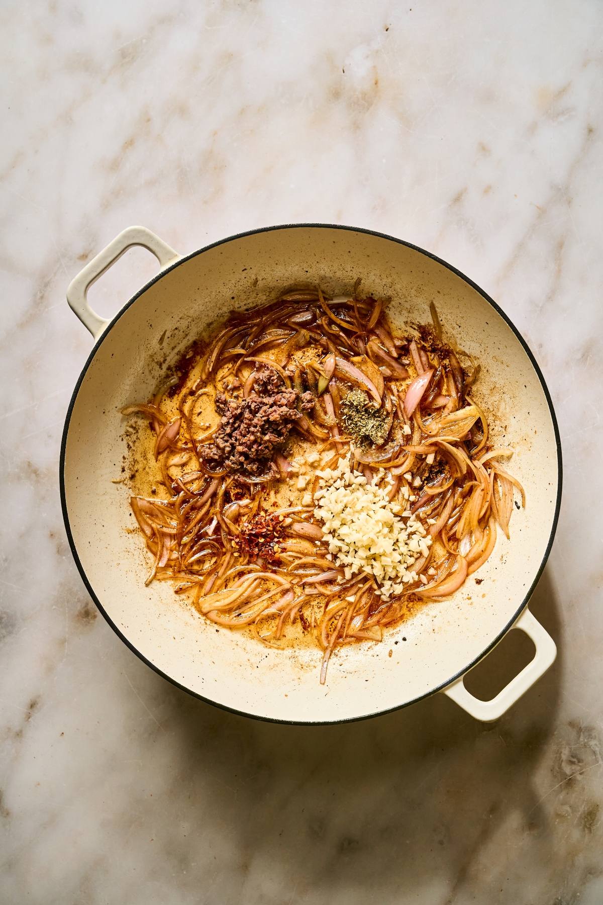 Sliced onions, minced garlic, and spices cooking in a large cream-colored pot on a marble countertop.