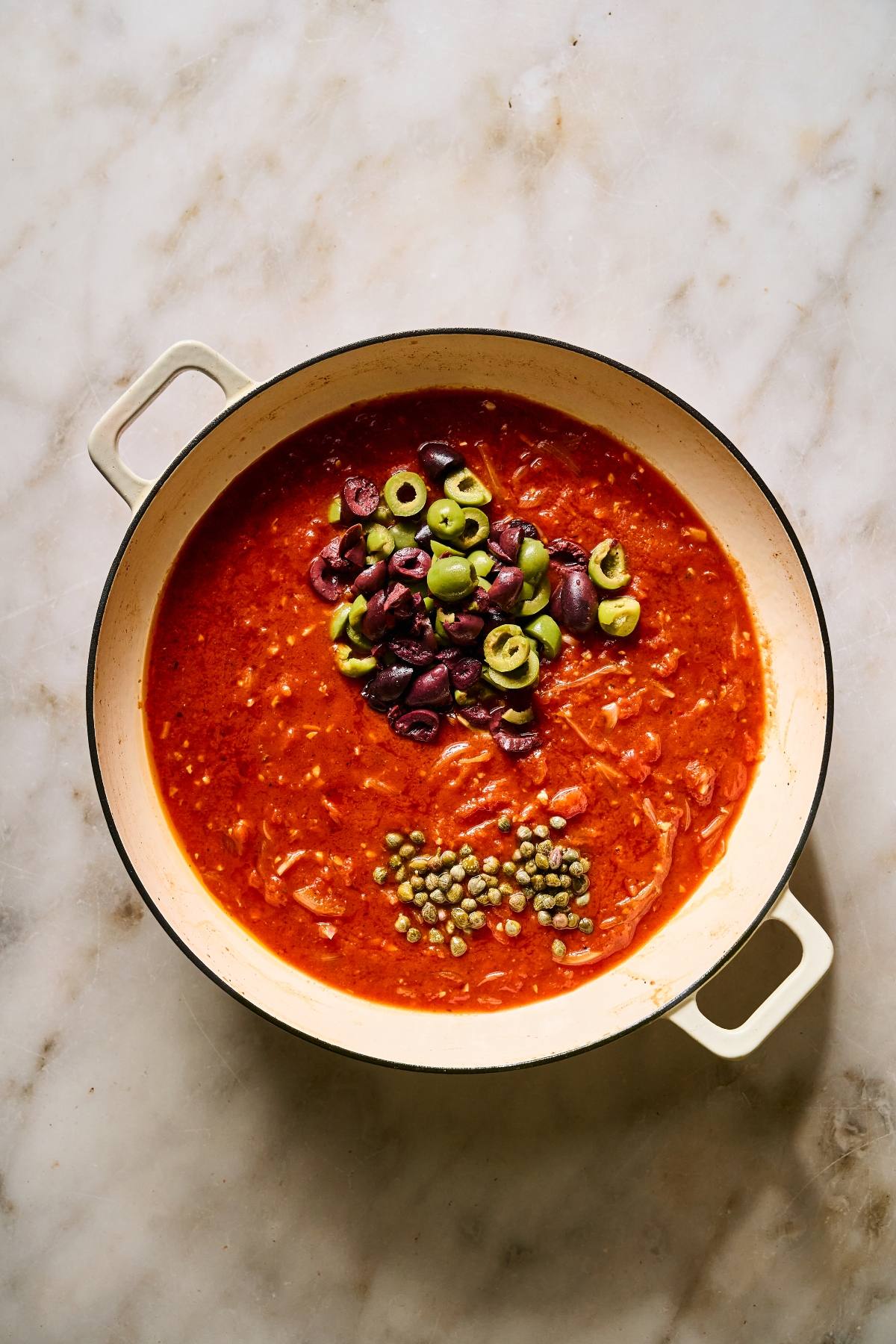 A pot of tomato sauce topped with sliced green and black olives and capers on a marble surface.