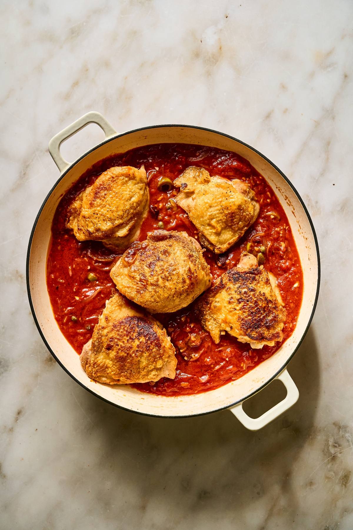 Four browned chicken thighs simmer in a red tomato sauce inside a round cream-colored Dutch oven.