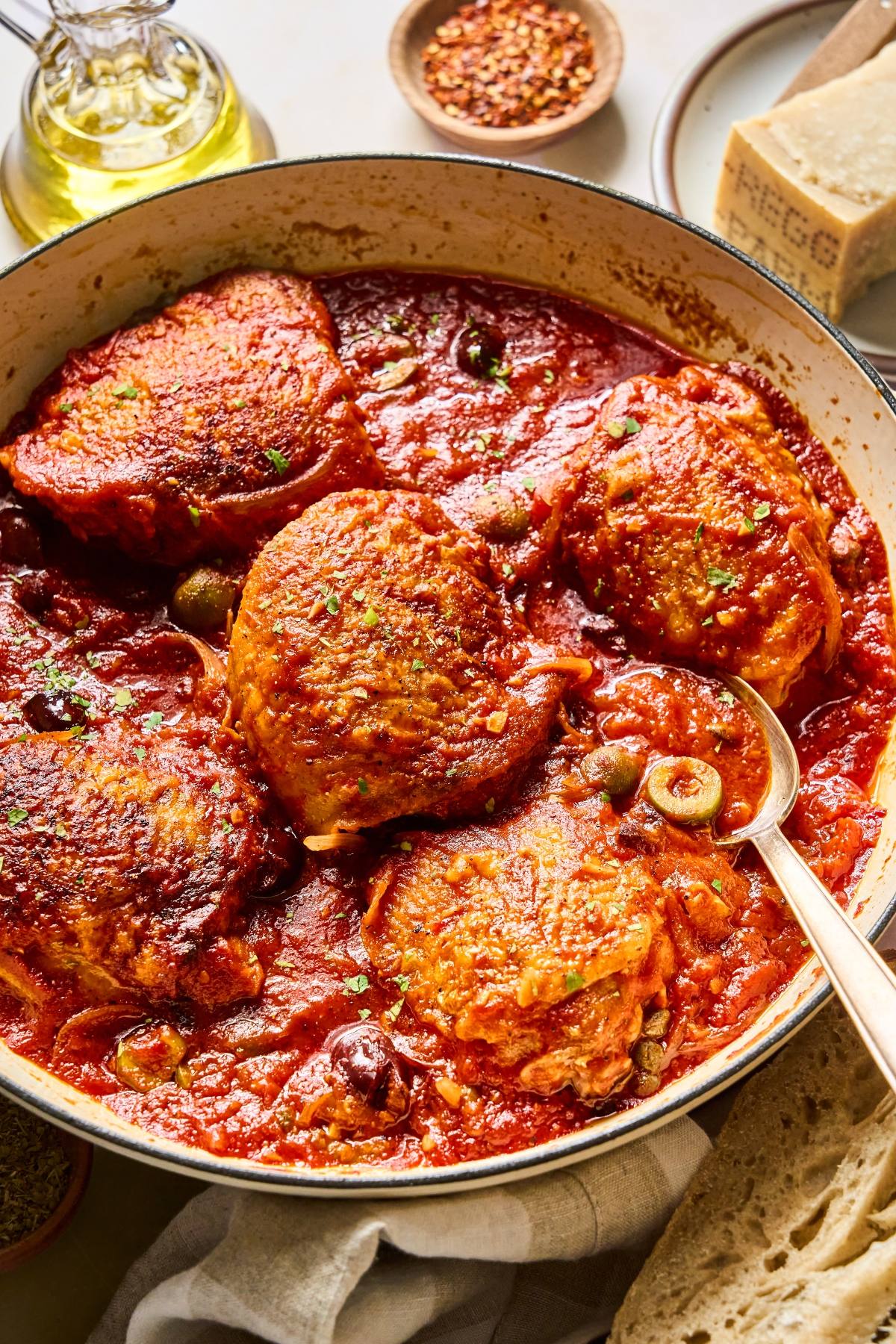 Golden brown chicken thighs simmer in a red tomato sauce with olives, served in a rustic pan with a spoon.