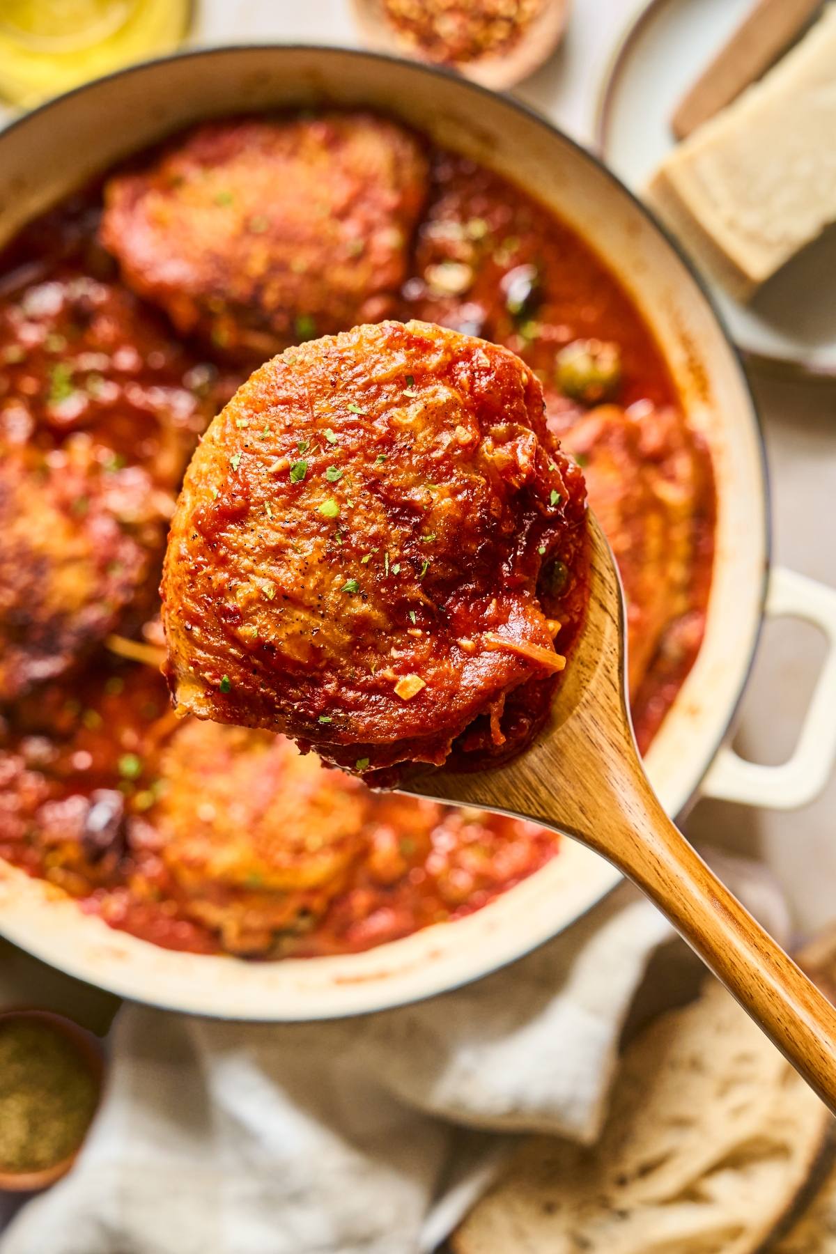 A wooden spoon holds a piece of chicken in tomato sauce over a pot with more chicken pieces.