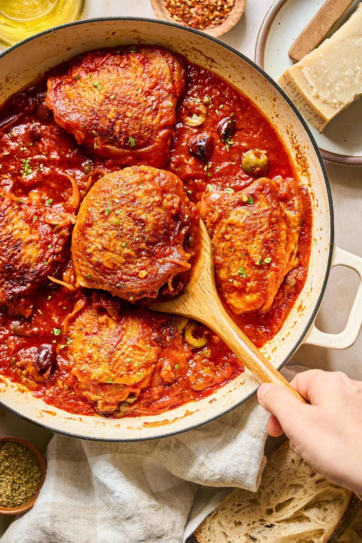 A hand serves braised chicken thighs in tomato sauce from a skillet, surrounded by bread and seasonings.