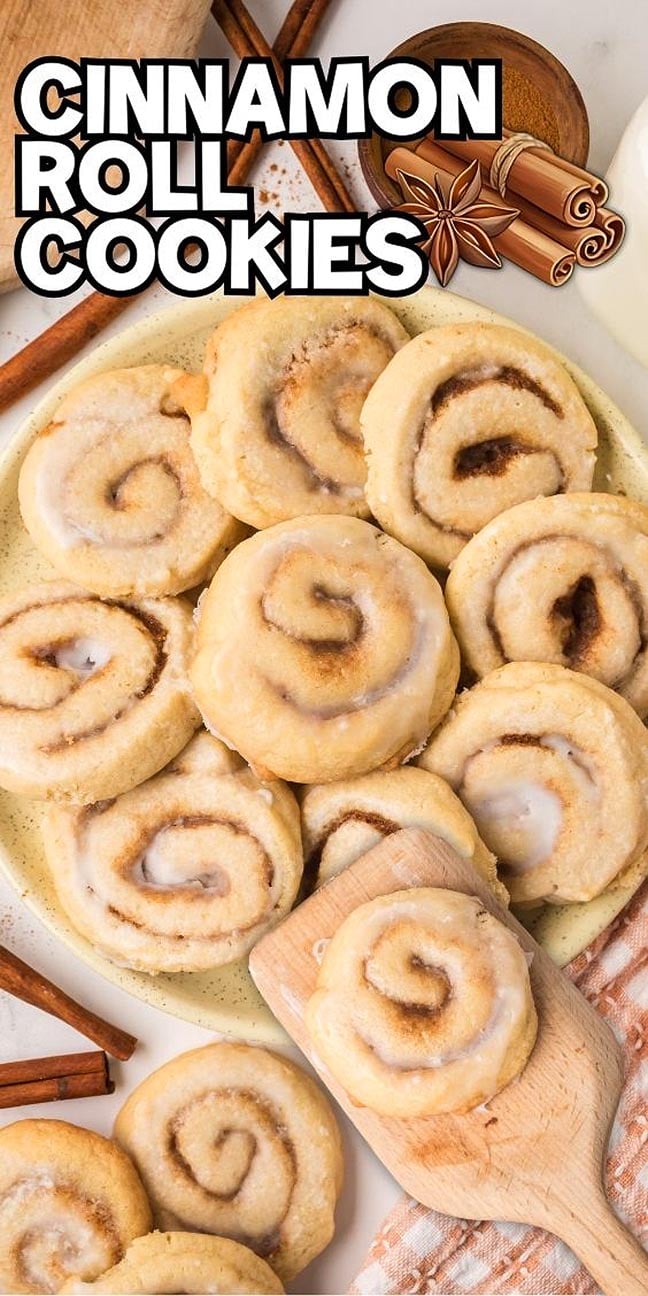 A plate of freshly baked Cinnamon Roll Cookies, perfectly glazed, sits beside a wooden spatula and aromatic cinnamon sticks.