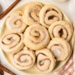 A plate of freshly baked Cinnamon Roll Cookies, frosted to perfection, sits on a table beside cinnamon sticks and a nearby jar.