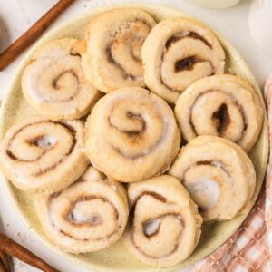 A plate of freshly baked Cinnamon Roll Cookies, frosted to perfection, sits on a table beside cinnamon sticks and a nearby jar.