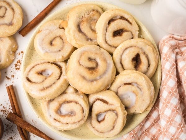 A plate of freshly baked Cinnamon Roll Cookies, frosted to perfection, sits on a table beside cinnamon sticks and a nearby jar.