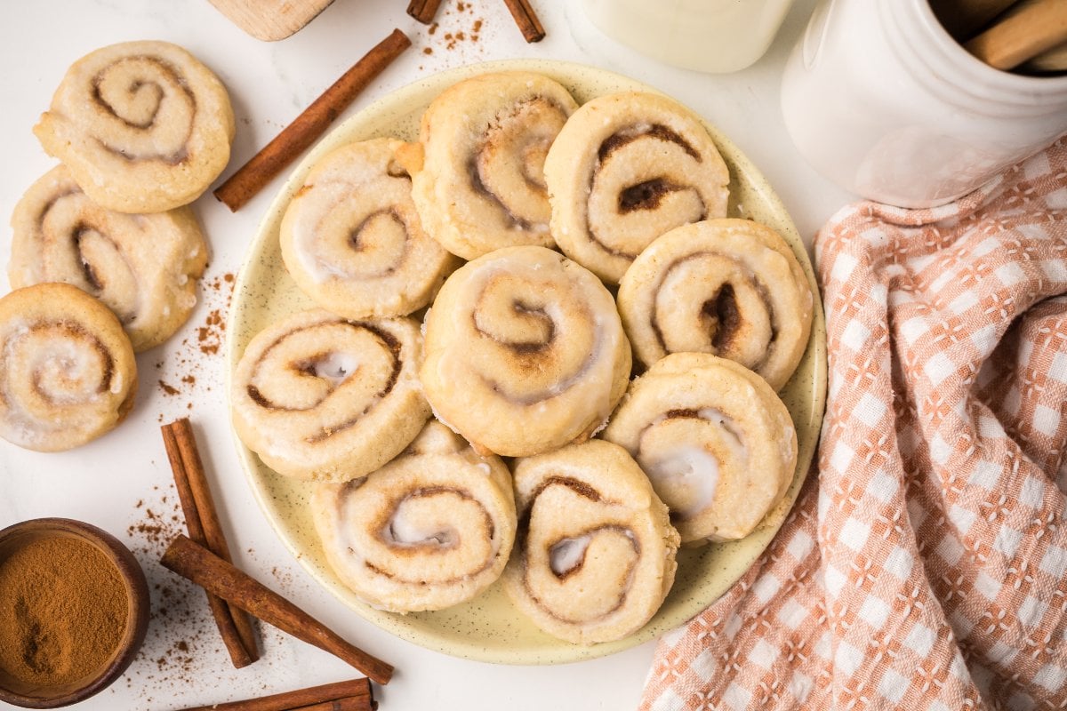 A plate of frosted Cinnamon Roll Cookies is surrounded by cinnamon sticks and a checked cloth napkin, creating a cozy and inviting dessert display.