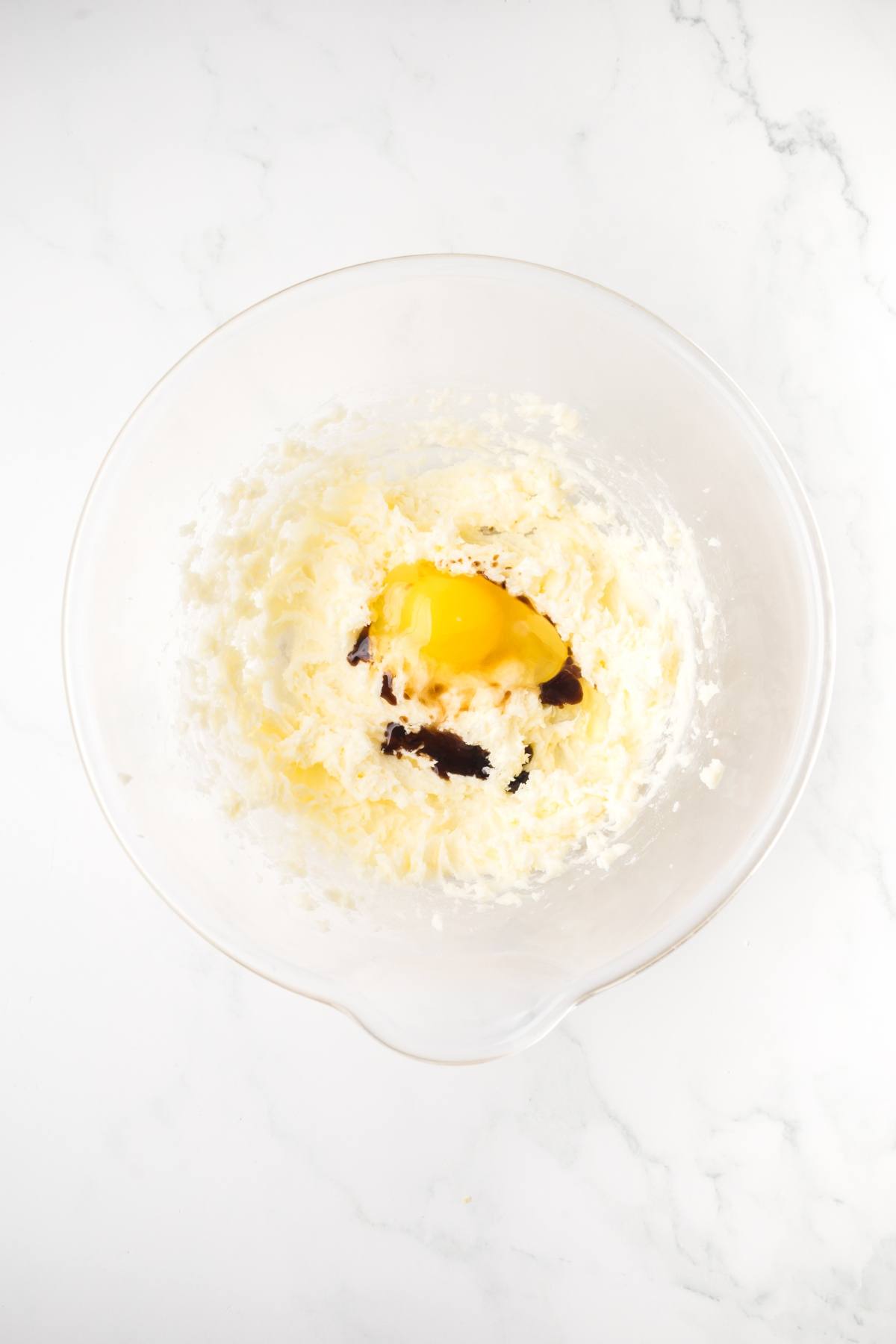 Clear mixing bowl with creamed butter, sugar, an egg, and vanilla extract on a white marble surface.