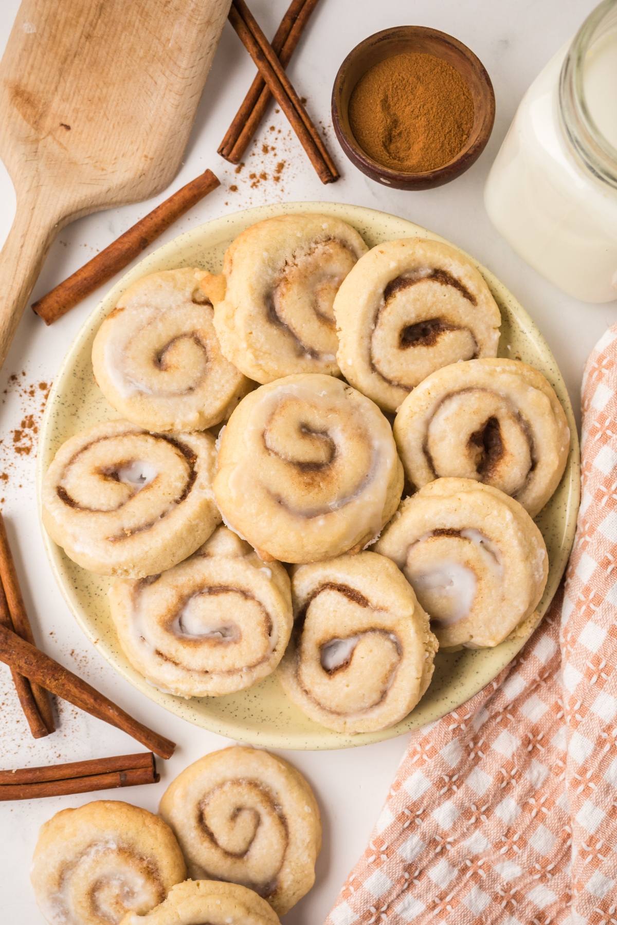 A plate of iced cinnamon rolls surrounded by cinnamon sticks, a jar of milk, and a small bowl of cinnamon.