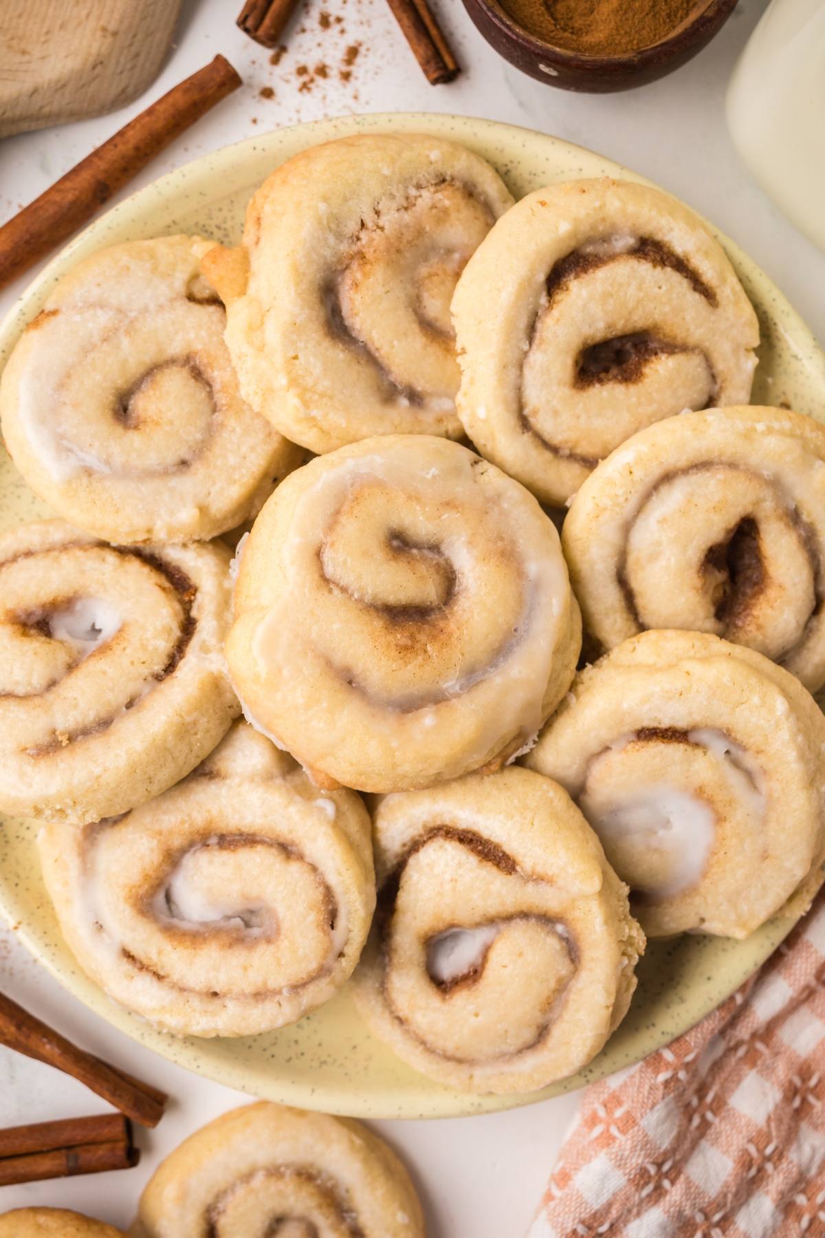 A plate of frosted cinnamon rolls is surrounded by cinnamon sticks and a checkered cloth.