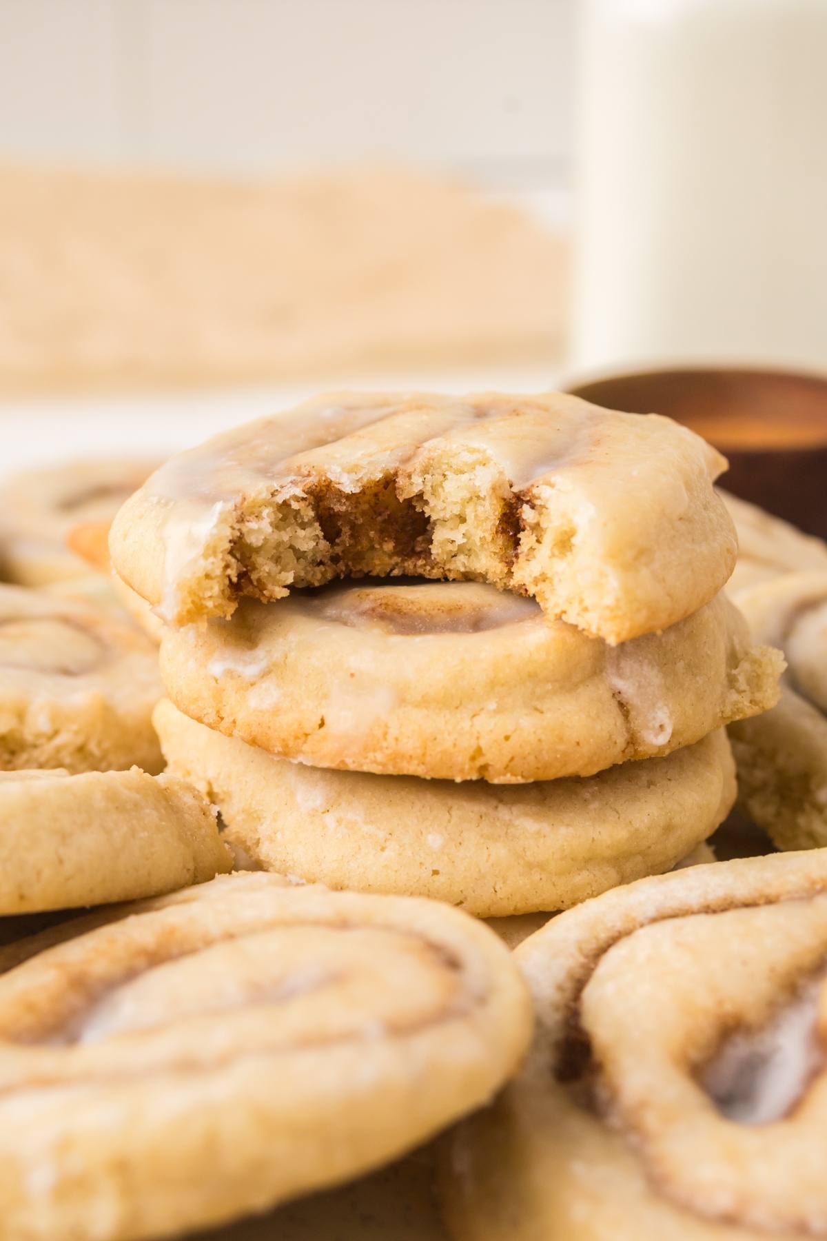 Two stacked cookies, one with a bite taken out, surrounded by more cookies and a glass of milk in the background.